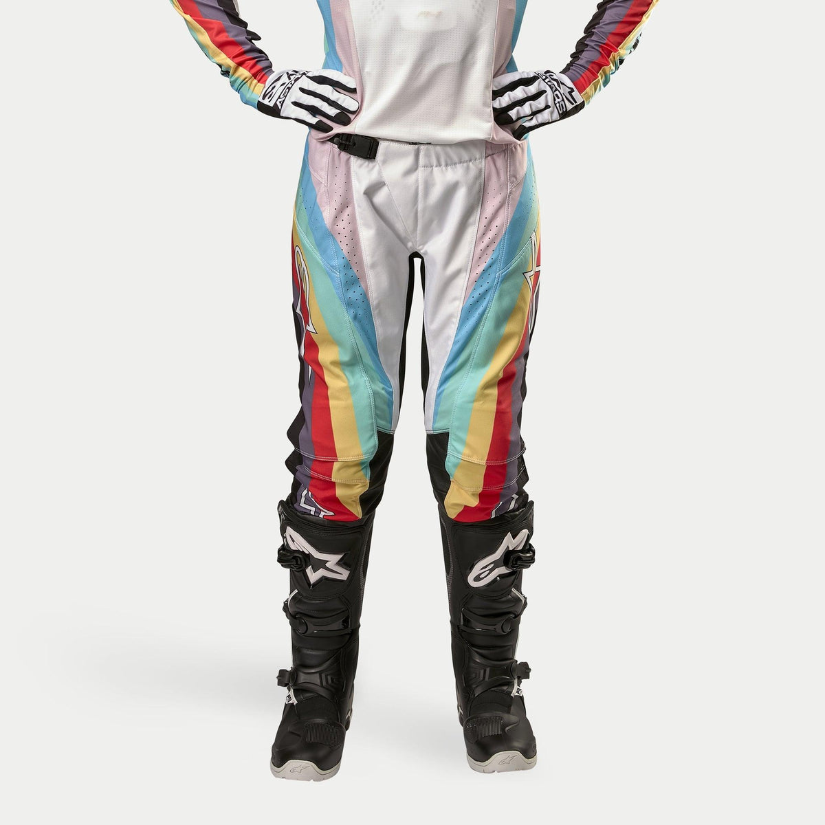 Alpinestars Stella Techstar Pants 2024 da donna, pantaloni da motocross, nero multicolore con vivaci strisce diagonali bianche, blu, rosse e gialle, indossati con stivali e guanti da motocross neri, design ispirato alle corse per prestazioni off-road.