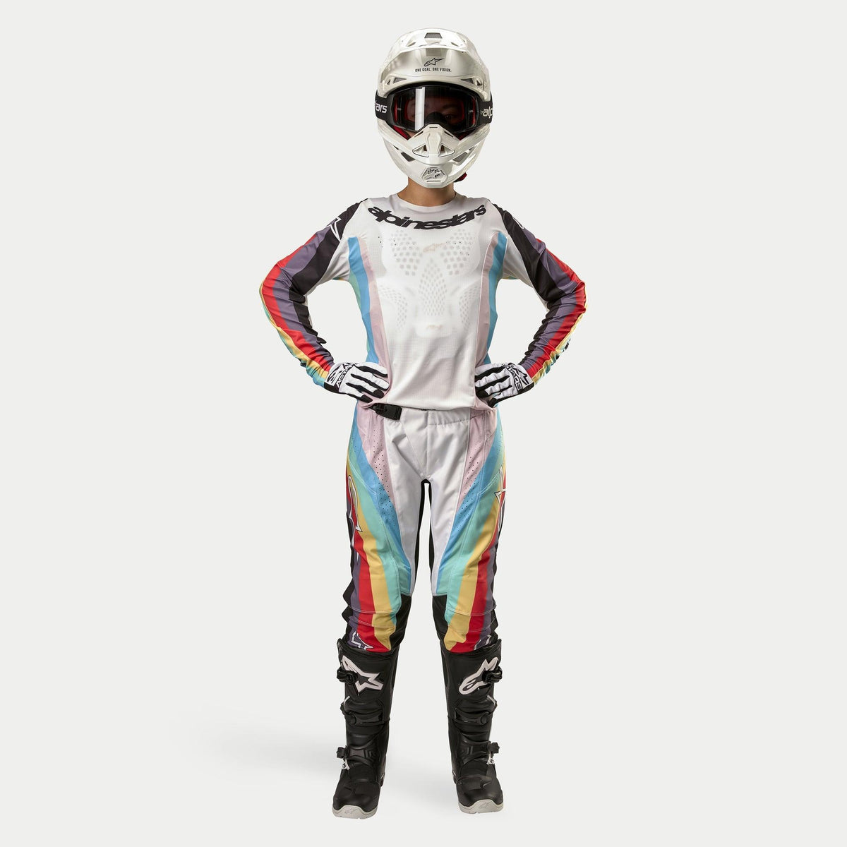 Pilota donna di motocross con pantaloni Alpinestars Stella Techstar 2024, Black Multicolor con base bianca e strisce rosse, gialle e blu, maglia a maniche lunghe coordinata, casco bianco e stivali neri, equipaggiamento da gara completo per fuoristrada