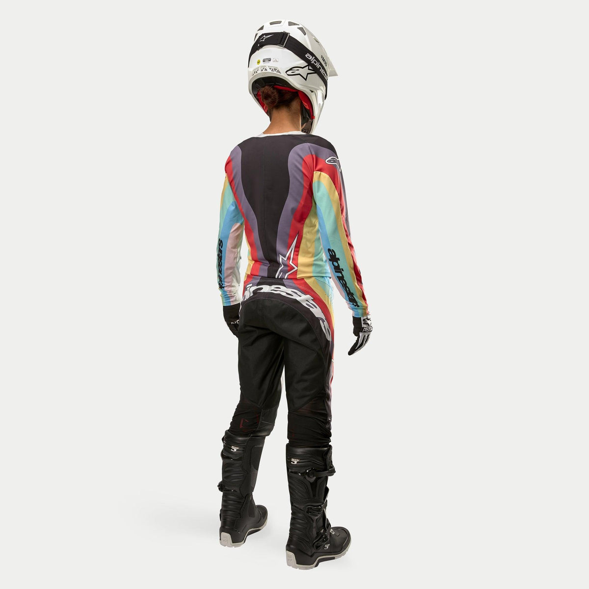 Pilota di motocross vista da dietro che indossa pantaloni Alpinestars Stella Techstar 2024 in Black Multicolor, con grande logo bianco sulla seduta, coordinati con una maglia a righe colorate, casco bianco e stivali Tech 3 neri per gare off-road.