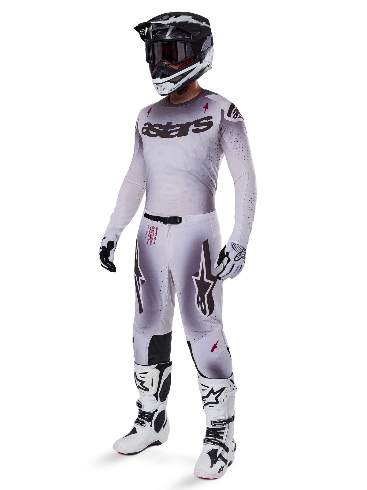 Pilota di motocross con abbigliamento completo da gara, maglia con sfumatura grigia e bianca con marchio 'astars' nero, pantaloni coordinati con dettagli rossi e loghi a stella, casco bianco e nero con maschera scura, stivali Alpinestars Tech 10 bianchi, completo professionale per fuoristrada