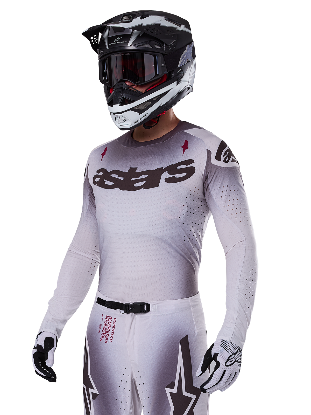 Pilota di motocross che indossa il set Alpinestars 2025 Supertech Maker in bianco, caratterizzato da una maglia con sfumatura grigia, logo astars nero e perforazioni tagliate al laser, pantaloni coordinati con fibbia magnetica e grafiche a stella, abbinato a casco e guanti bianchi e neri per un look completo da gara fuoristrada.