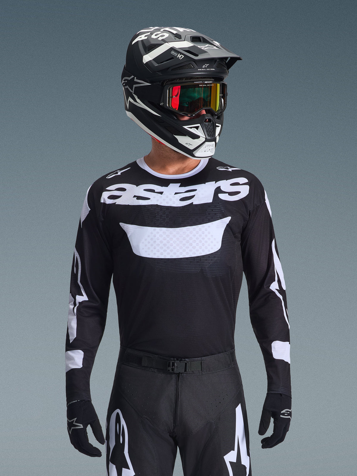 Pilota di motocross che indossa il completo Alpinestars 2026 Racer Air Riway, maglia nera con logo 'astars' bianco e pannello ventilato sul petto, pantaloni neri con accenti bianchi, casco coordinato nero e bianco con occhiali iridescenti, guanti neri, abbigliamento da motocross ad alte prestazioni in nero e bianco.