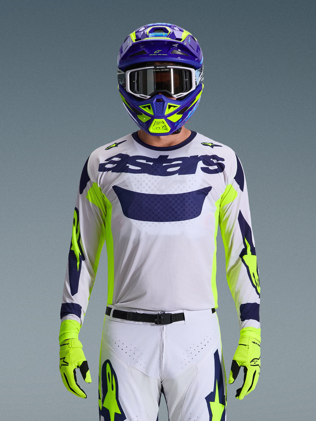 Maglia Alpinestars Racer Air Riway 2026, grigio chiaro con logo astars blu scuro, accenti giallo fluo e blu sulle maniche, indossata da un pilota di motocross con casco e guanti coordinati viola e giallo neon, tessuto tecnico in rete per gare off-road
