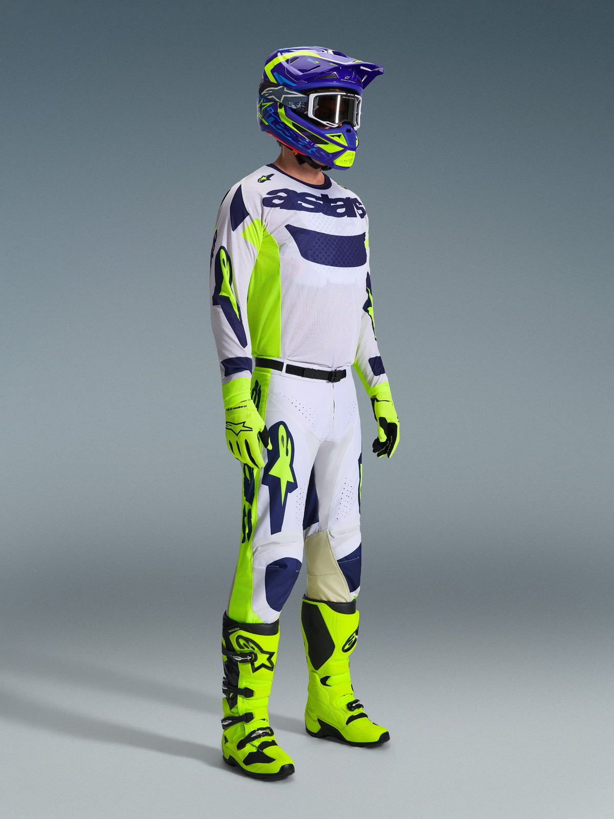 Pilota di motocross con abbigliamento da gara completo, maglia grigio chiaro con accenti giallo fluo e blu, pantaloni coordinati bianchi e gialli, stivali e guanti giallo acceso, casco viola e giallo con occhiali scuri, completo coordinato per corse fuoristrada