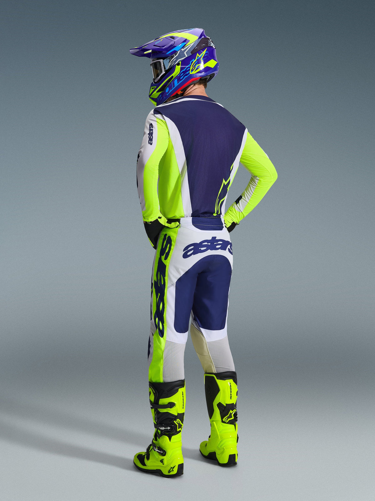 Pilota di motocross Alpinestars visto da dietro che indossa la maglia Racer Air Riway 2026 in grigio chiaro, giallo fluo e blu, pantaloni coordinati con logo evidente, stivali da motocross giallo fluo e casco blu e giallo, set completo da gara off-road