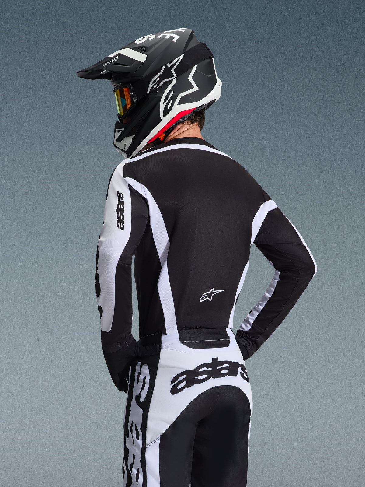 Pilota di motocross visto da dietro che indossa la maglia Alpinestars 2026 Racer Riway in bianco e nero, con marchio 'astars' sulle maniche e sulla parte bassa della schiena, abbinata a pantaloni da motocross coordinati e casco nero con occhiali, equipaggiamento coordinato per gare off-road.