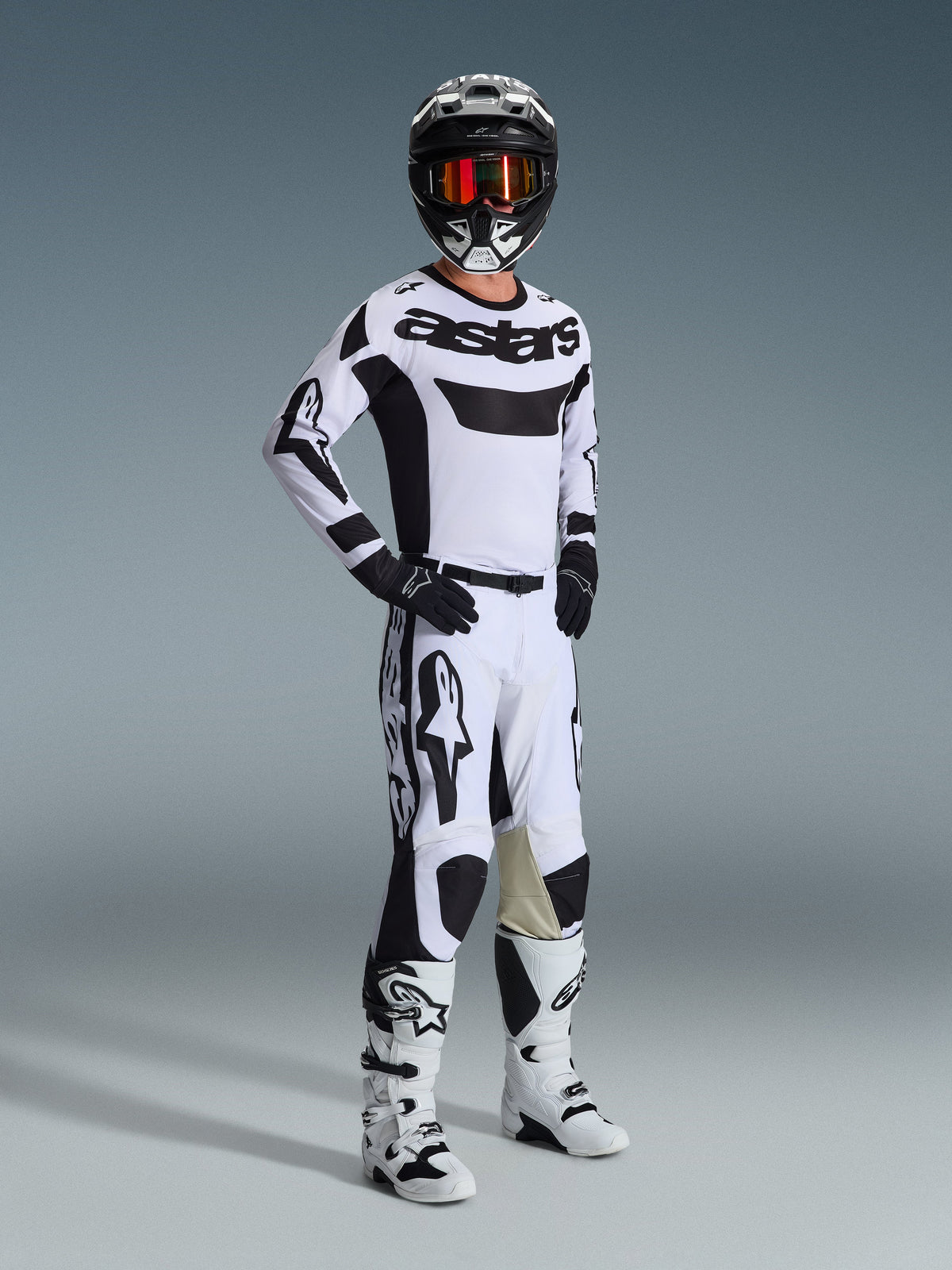 Pilota di motocross con abbigliamento completo da gara, maglia Alpinestars 2026 Racer Riway in bianco e nero, pantaloni da motocross coordinati bianchi e neri, casco nero con occhiali a specchio, stivali bianchi e neri e guanti neri, outfit coordinato per competizioni off-road