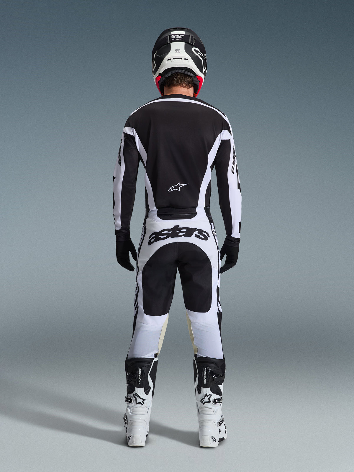 Vista posteriore di un pilota di motocross con il completo 2026 Racer Riway in bianco e nero, caratterizzato da maglia nera con accenti bianchi, pantaloni bianchi con vistoso marchio 'astars' sulla seduta, casco nero, stivali bianchi e guanti neri, abbigliamento professionale completo da gara off-road.