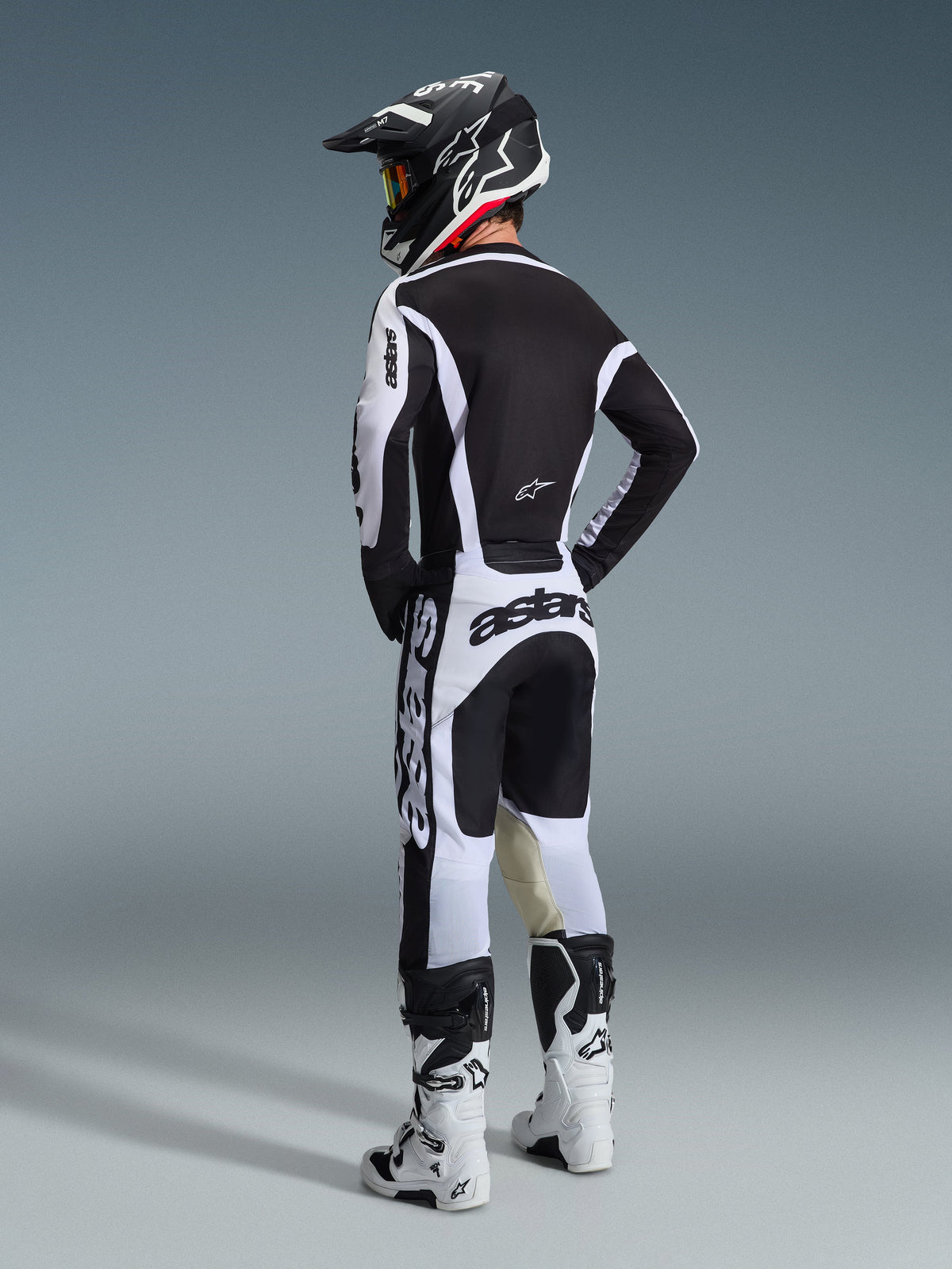 Pilota di motocross con abbigliamento completo da gara, vista posteriore, maglia Alpinestars 2026 Racer Riway bianca e nera, pantaloni coordinati bianchi e neri con vistoso marchio 'astars', stivali bianchi e casco nero con accenti bianchi, outfit tecnico per corse fuoristrada