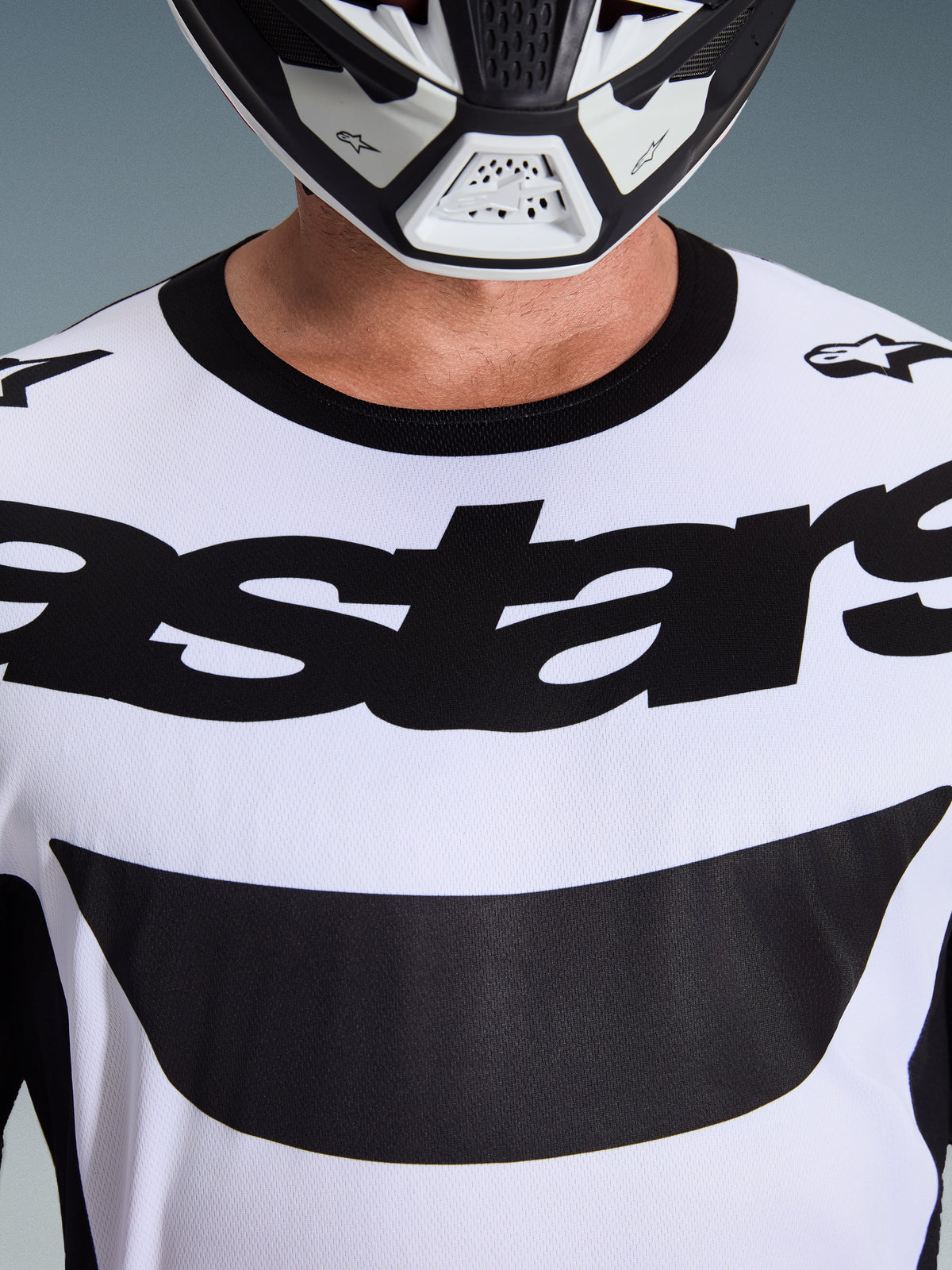 Alpinestars Maglia Racer Riway 2026, maglia da motocross, bianco e nero, primo piano del petto con vistoso logo astars nero e colletto nero, indossata con casco bianco e nero, abbigliamento tecnico per fuoristrada