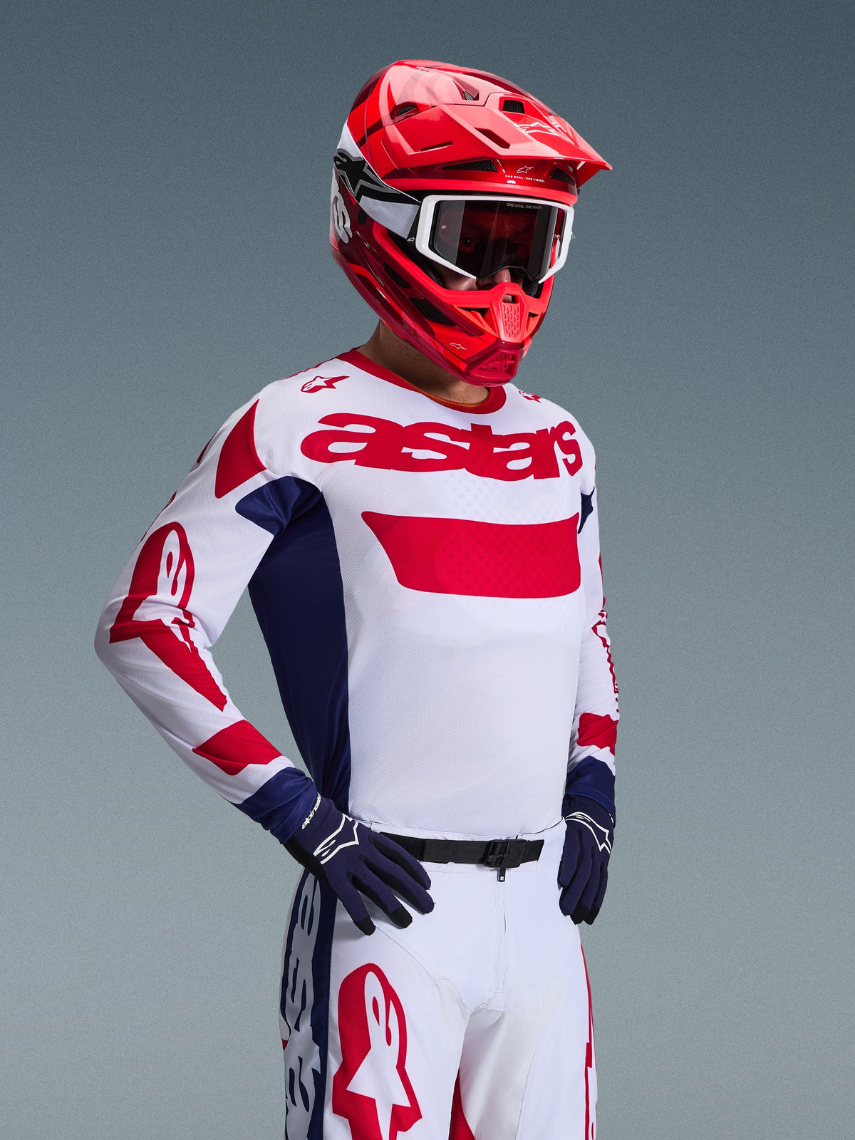 Pilota di motocross che indossa la maglia Alpinestars Racer Riway 2026 in bianco, blu e rosso, pantaloni da fuoristrada coordinati, casco rosso acceso con maschera bianca e guanti blu navy, completo tecnico ad alte prestazioni coordinato per il motocross