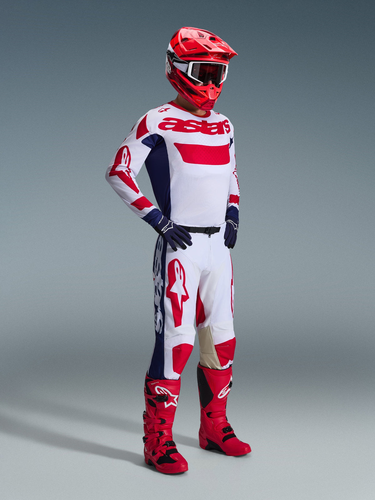 Pilota di motocross con completo Alpinestars 2026 Racer Riway, maglia bianca con accenti rossi e blu e branding 'astars', pantaloni bianchi coordinati con grandi loghi rossi e pannelli laterali blu navy, casco rosso, stivali rossi e neri e guanti blu navy, kit da gara professionale per fuoristrada.