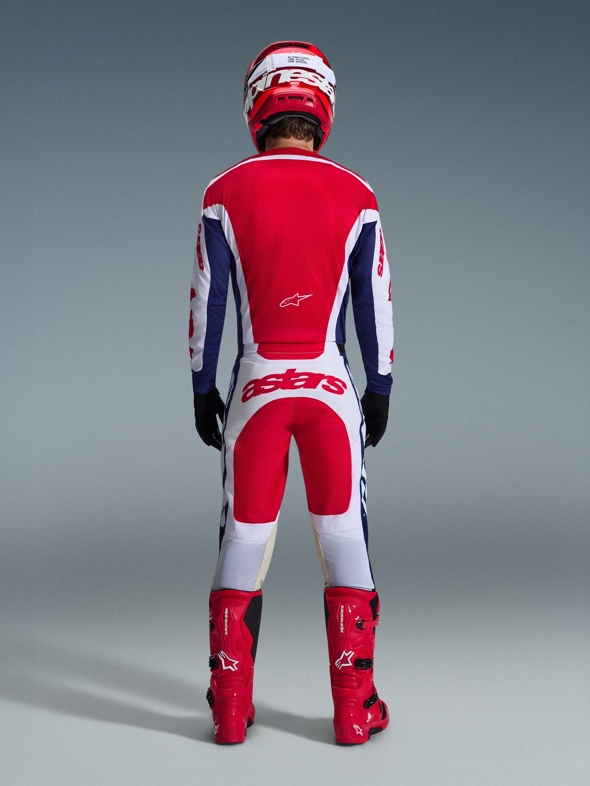 Pilota di motocross visto da dietro che indossa il completo Racer Riway 2026, maglia rossa e blu con accenti bianchi, pantaloni bianchi con pannelli rossi e logo 'astars' sulla seduta, coordinato con casco rosso, stivali rossi e guanti neri, abbigliamento da gara ad alte prestazioni per il fuoristrada