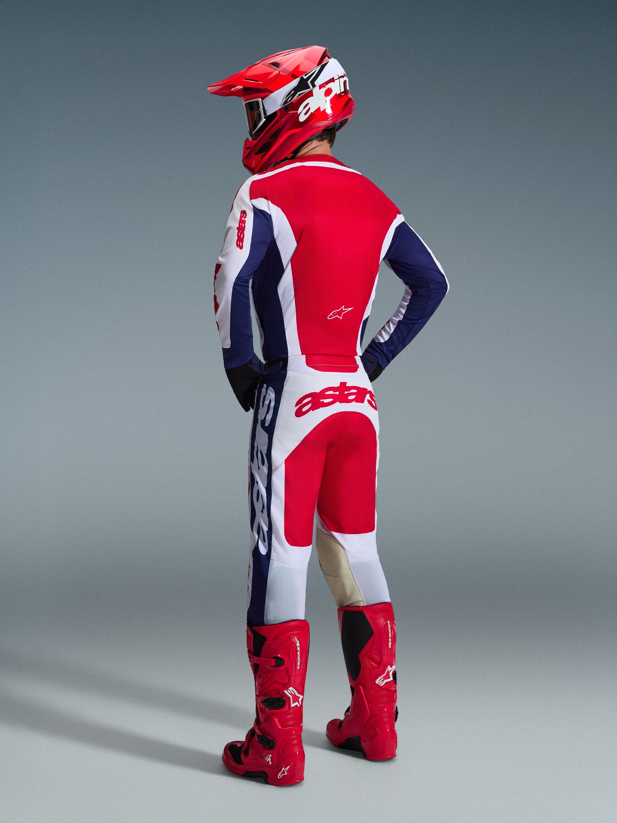 Pilota di motocross visto da dietro che indossa il completo Alpinestars 2026 Racer Riway in Bianco Blu Rosso, inclusa una maglia a maniche lunghe con marchio Astars e pantaloni coordinati, abbinati a un casco rosso e stivali da fuoristrada rossi.