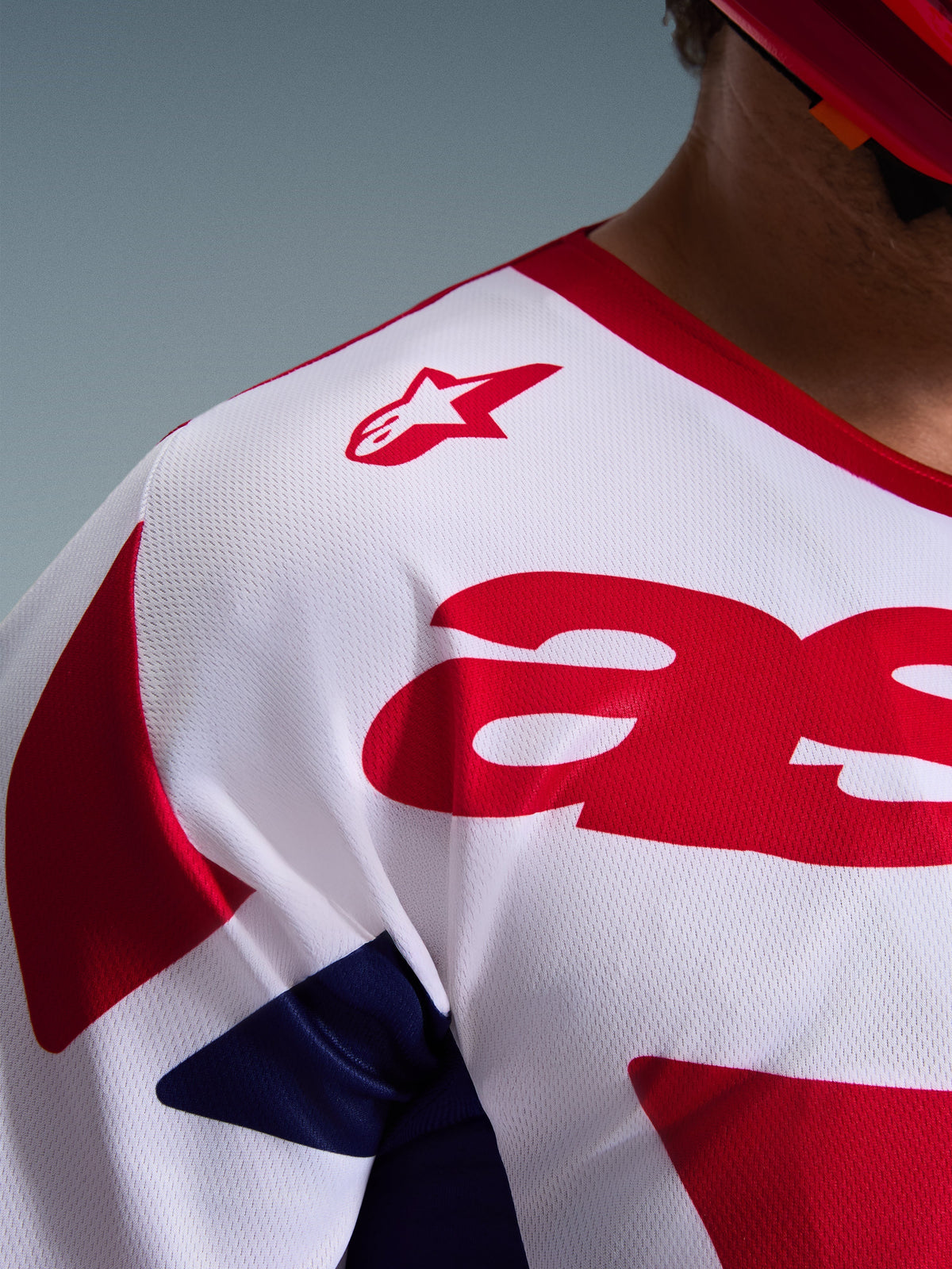 Dettaglio ravvicinato della maglia Alpinestars Racer Riway 2026 in bianco, blu e rosso, che mette in risalto la struttura in poli-tessuto a rete traspirante, il logo Alpinestars rosso sulla spalla e il materiale anti-umidità per le corse fuoristrada.