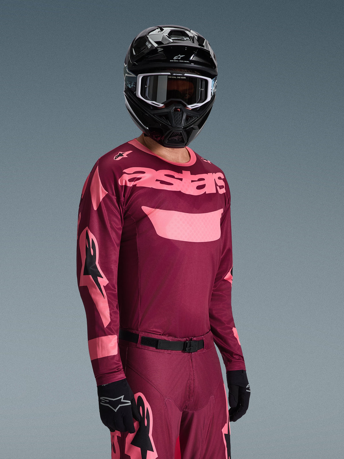 Pilota di motocross che indossa il completo Alpinestars Racer Riway 2026, maglia bordeaux e rosa con logo 'astars' in evidenza, pantaloni coordinati con dettagli rosa, casco e guanti neri, abbigliamento da gara professionale per motocross off-road