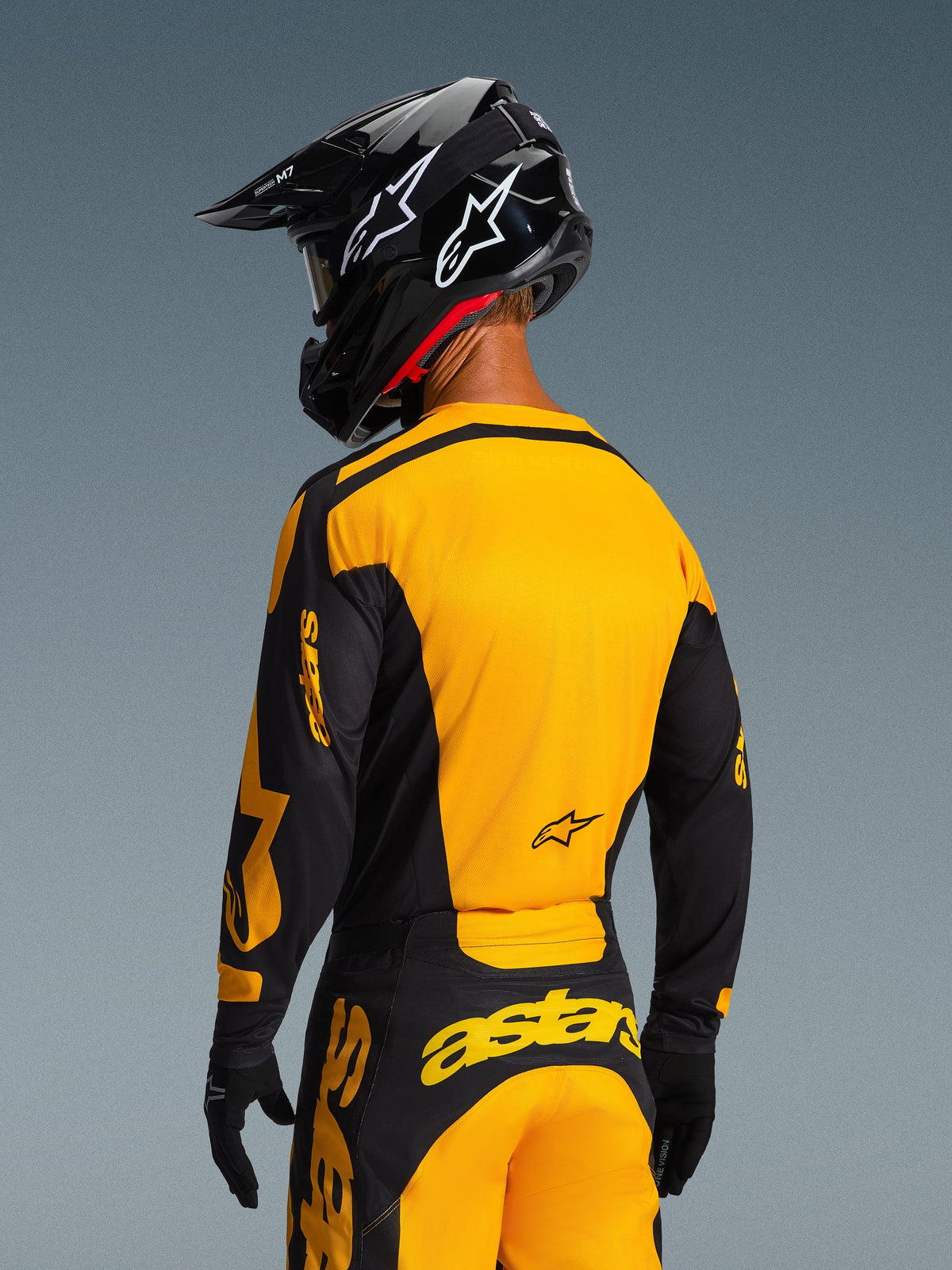 Pilota di motocross visto da dietro che indossa la maglia Racer Riway 2026 e i pantaloni coordinati in giallo e nero, con evidente marchio Alpinestars, casco nero con loghi a stella bianchi e guanti neri, set completo da motocross per corse off-road.