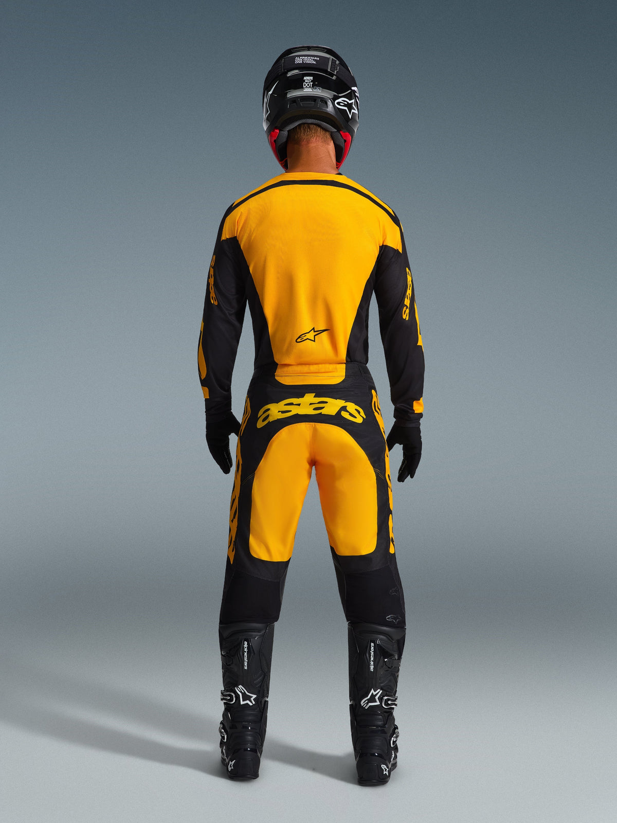 Pilota di motocross visto da dietro con maglia Alpinestars Racer Riway 2026 e pantaloni coordinati giallo e nero, casco nero, stivali e guanti neri, kit completo da corsa off-road con marchio Alpinestars in evidenza