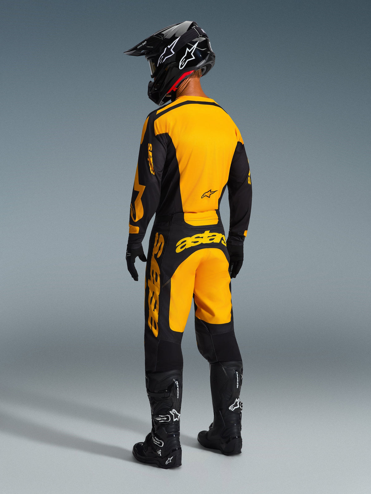 Pilota di motocross visto da dietro che indossa il completo Alpinestars Racer Riway 2026, maglia gialla e nera con pantaloni abbinati, casco nero, guanti neri e stivali neri con dettagli bianchi, outfit coordinato per corse off-road professionali.
