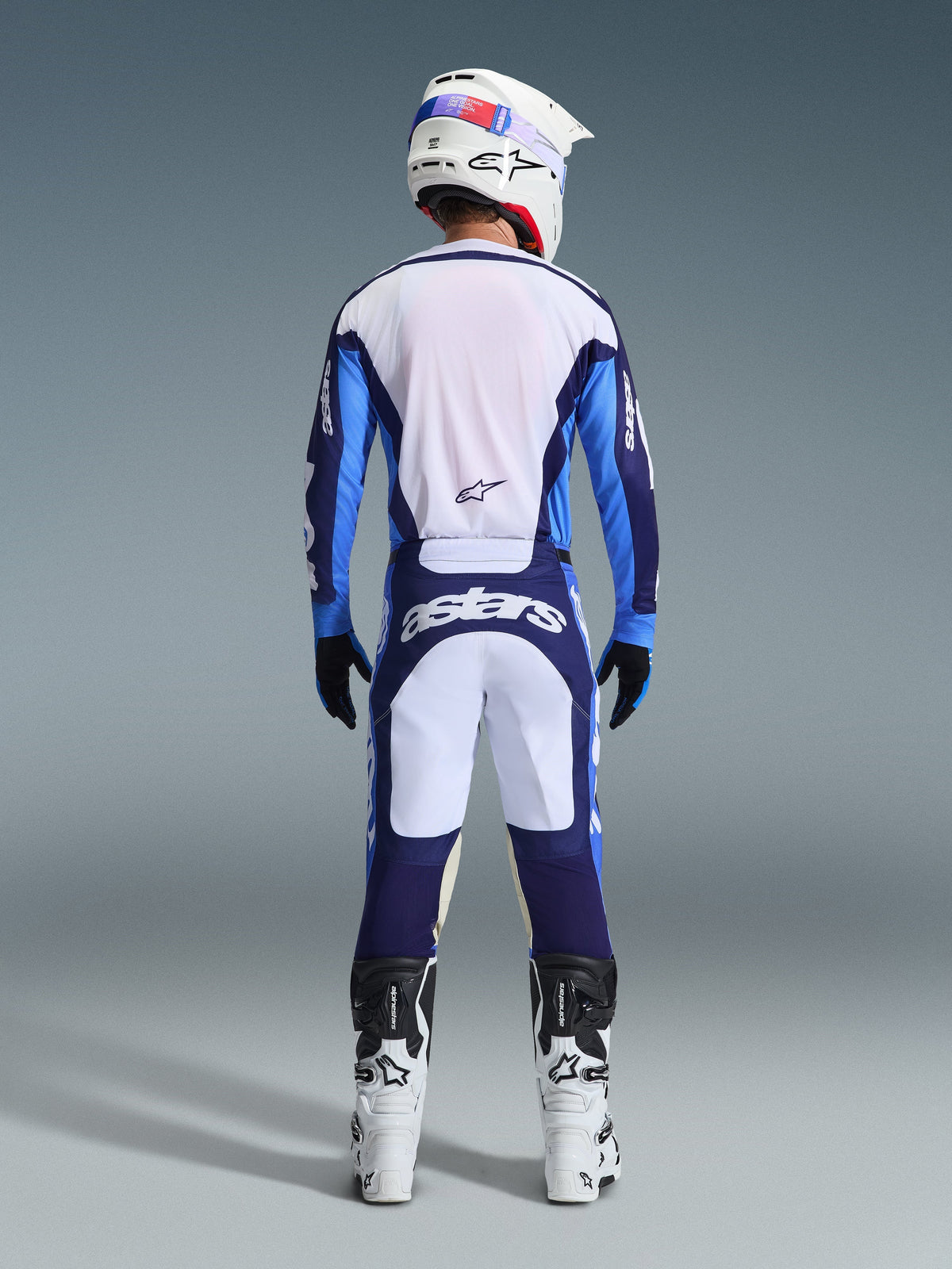 Vista posteriore di un pilota di motocross che indossa la maglia e i pantaloni Alpinestars 2026 Racer Riway in blu Ucla e bianco, coordinati con casco bianco, guanti neri e stivali da motocross bianchi, equipaggiamento ad alte prestazioni per competizioni off-road