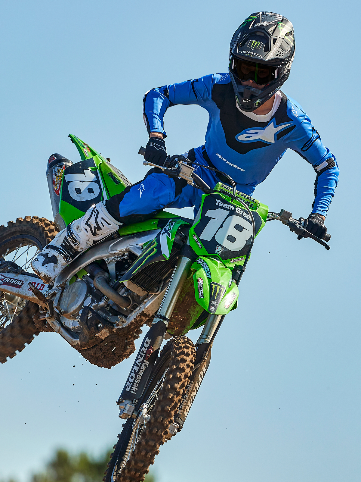 Alpinestars 2025 Racer Veil Jersey, maglia da motocross, blu e nera, maglia tecnica a maniche lunghe con grafiche astratte e sezioni in rete traspirante, indossata da un pilota durante un salto off-road su una moto verde