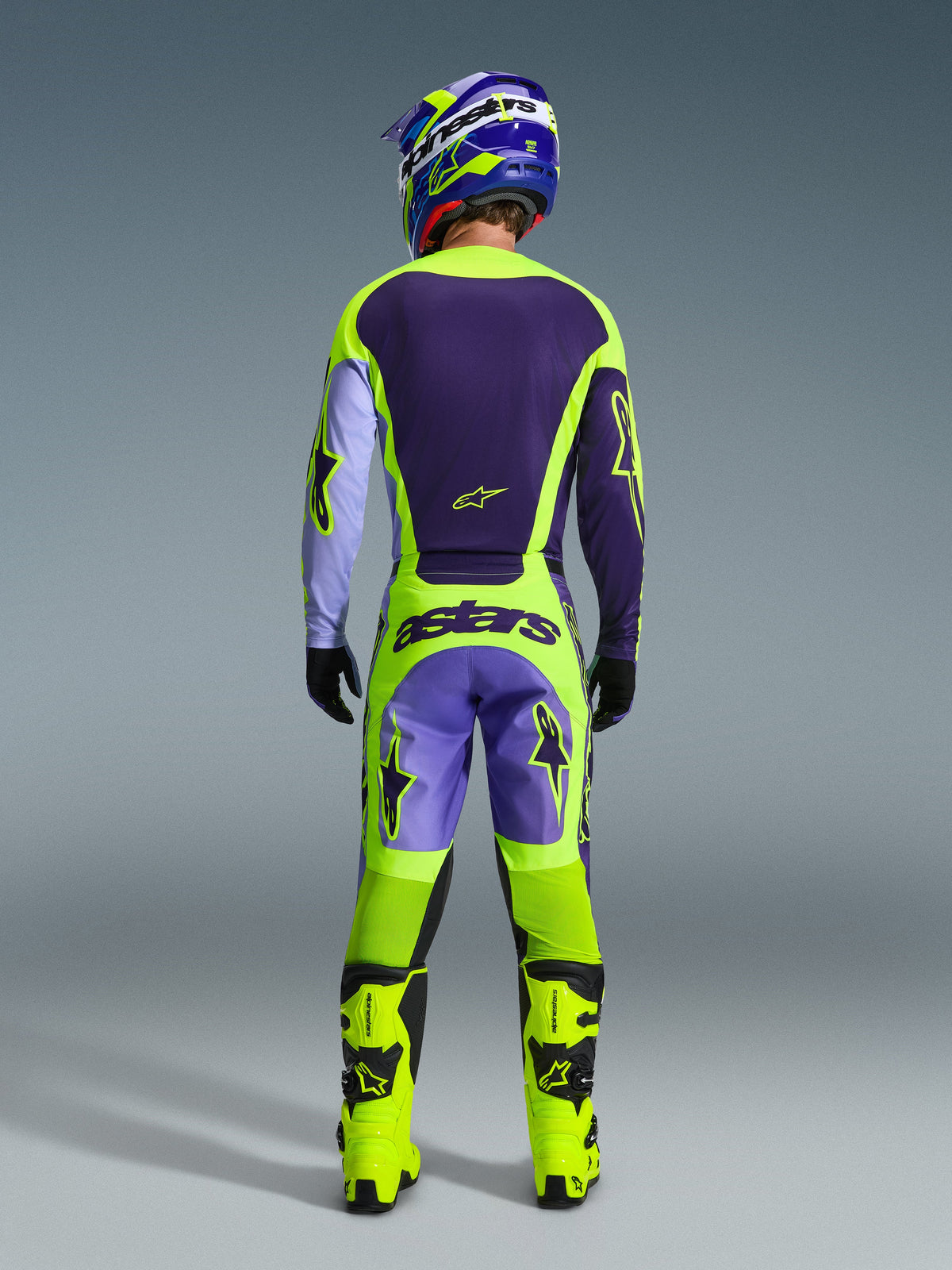 Pilota di motocross con completo Alpinestars 2026 Racer Portl, vista posteriore, maglia viola scuro e giallo neon con maniche color lavanda, pantaloni coordinati con branding 'astars', stivali giallo neon vivace, abbigliamento professionale da gara fuoristrada