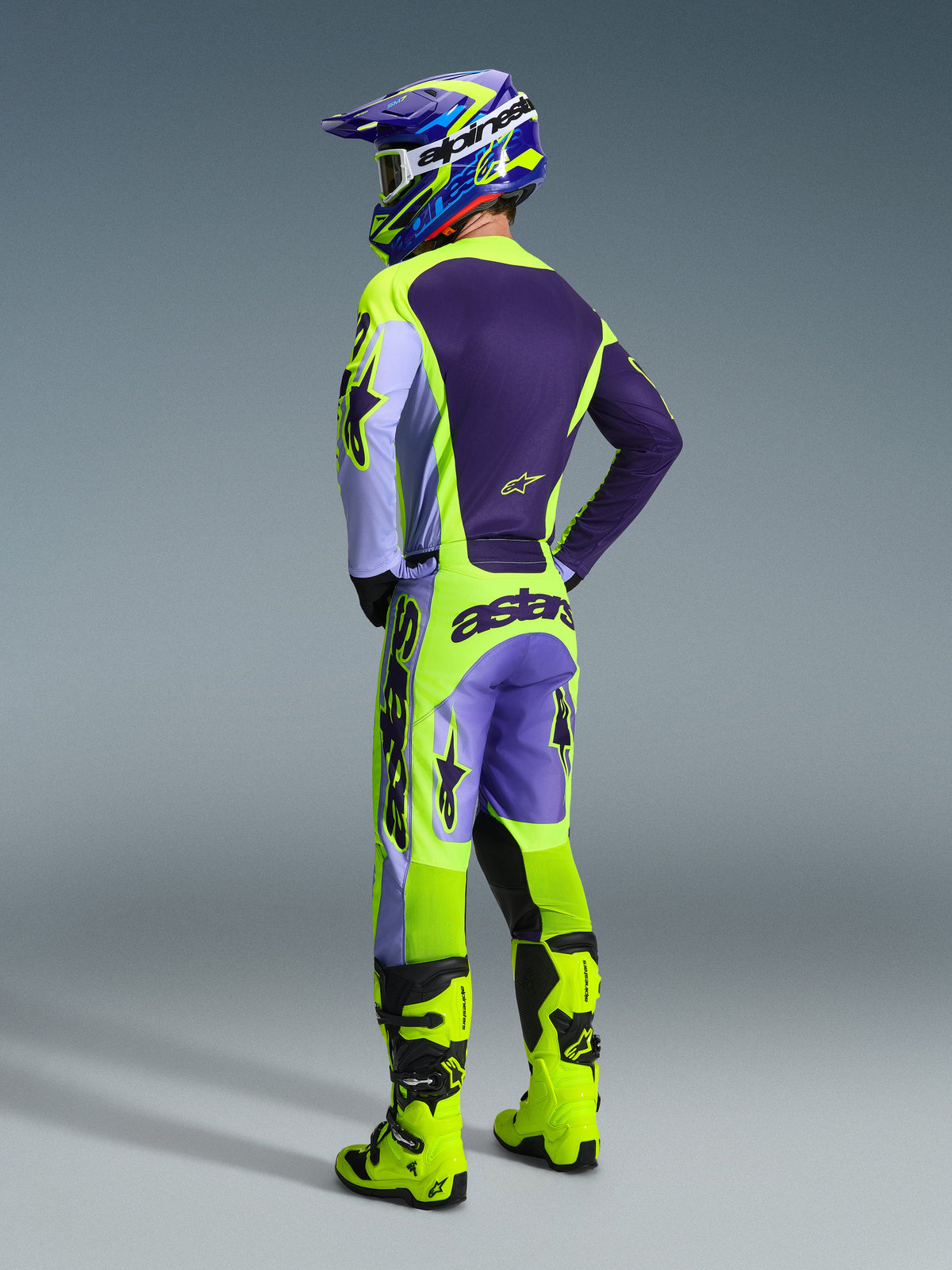 Pilota di motocross che indossa il completo Alpinestars Racer Portl 2026, maglia e pantaloni viola e giallo neon con pannelli lavanda, casco coordinato viola e giallo, stivali giallo neon brillante, vista posteriore dell'equipaggiamento professionale da corsa off-road.