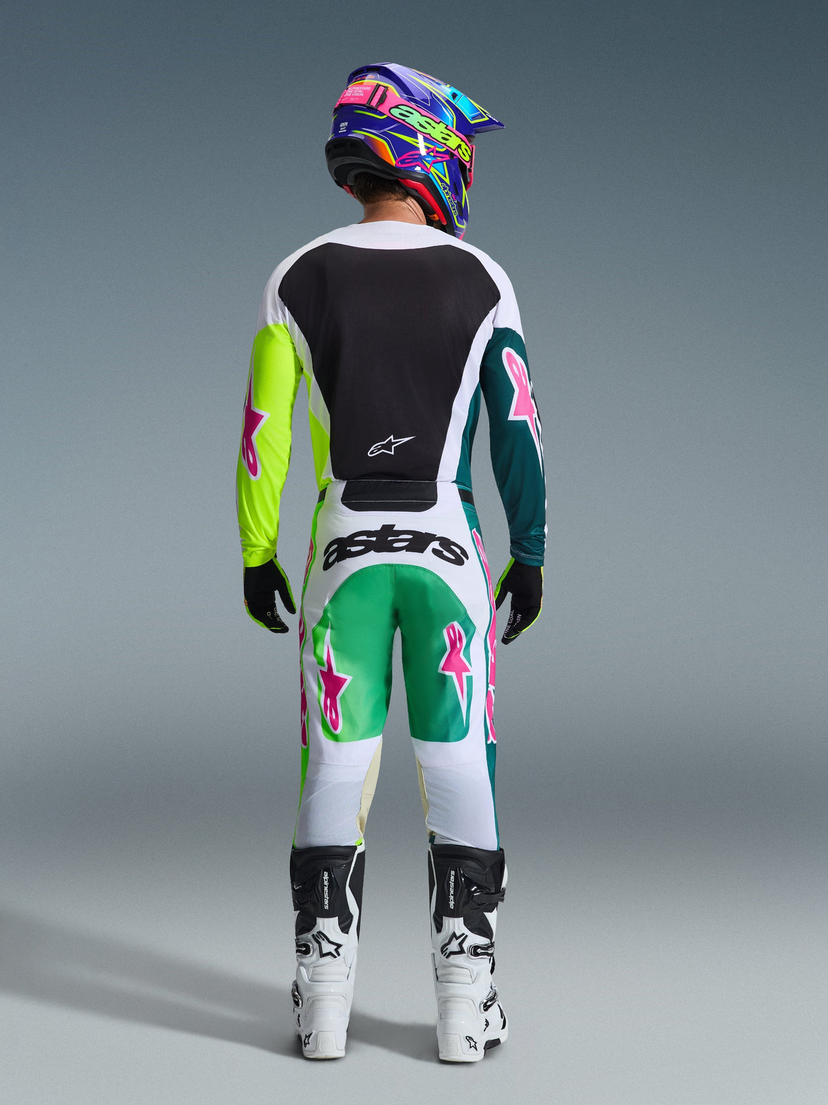 Pilota di motocross visto di spalle che indossa la maglia Alpinestars Racer Portl 2026 nella colorazione Green White Black, con pannello posteriore nero e maniche asimmetriche verdi con loghi a stella rosa, abbinata a pantaloni da motocross coordinati e stivali bianchi.