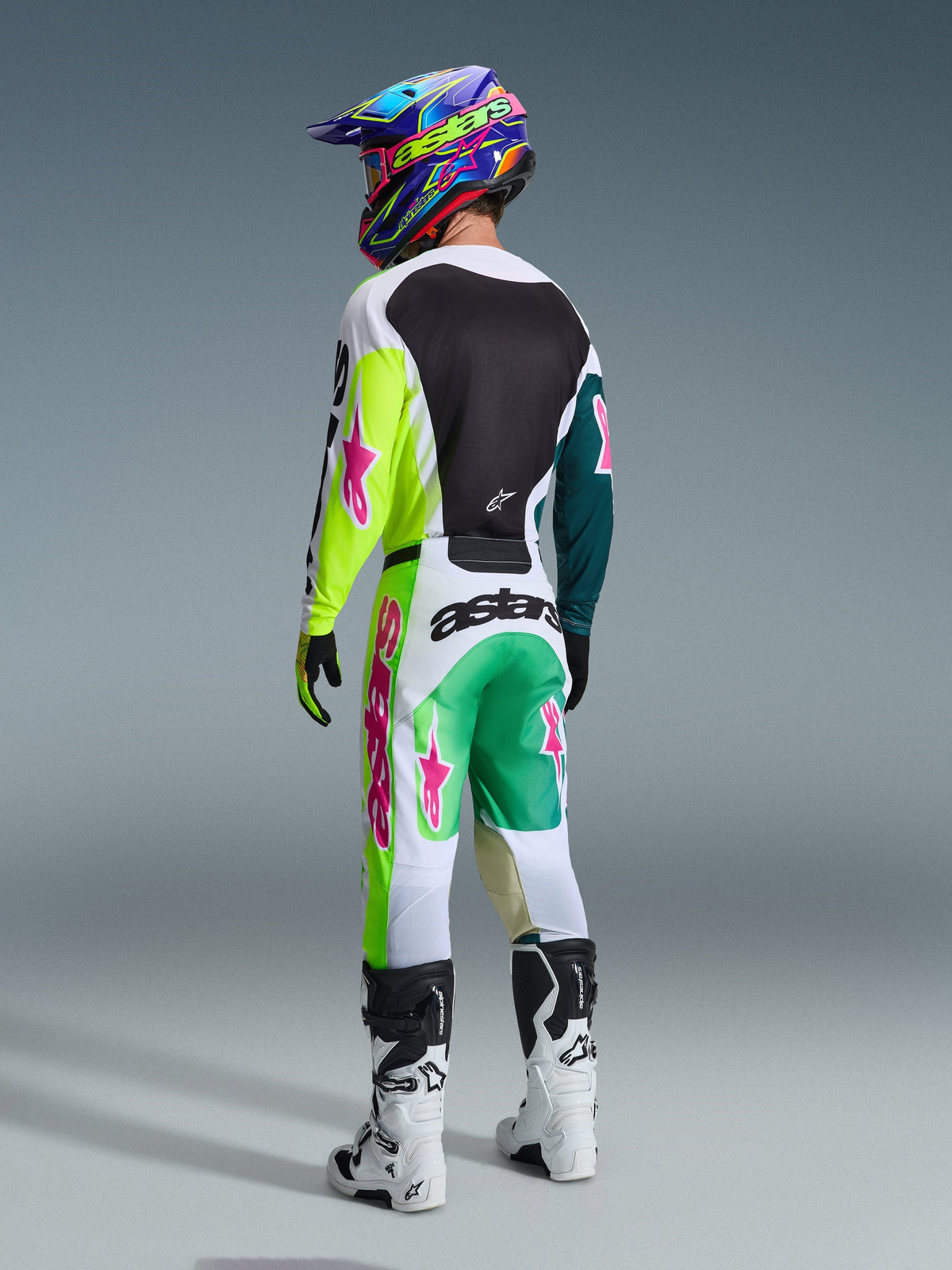 Pilota di motocross che indossa la maglia Alpinestars 2026 Racer Portl in verde, bianco e nero, visto da dietro, pantaloni da gara coordinati con loghi a stella rosa e marchio 'astars', stivali bianchi e neri e un casco colorato, outfit completo e coordinato per corse fuoristrada