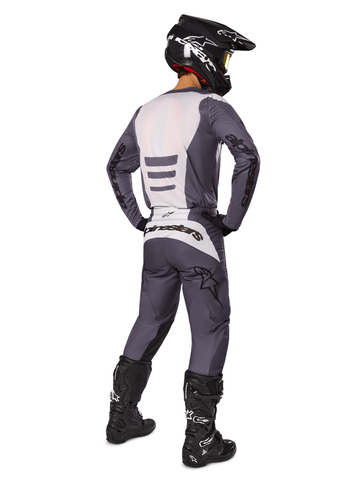 Pilota di motocross visto da dietro che indossa il set di abbigliamento Alpinestars 2025 Fluid Haul, maglia grigia e bianca e pantaloni coordinati, casco nero con occhiali gialli, stivali da motocross neri, equipaggiamento da gara completo per prestazioni fuoristrada
