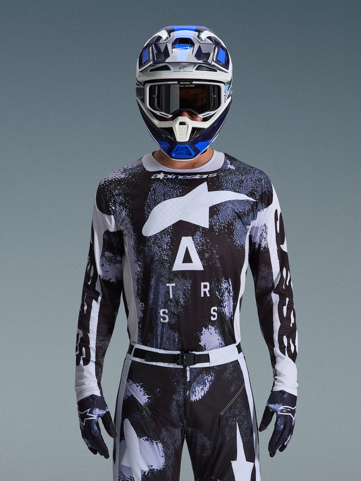 Pilota di motocross con il completo Alpinestars 2026 Racer Lahnd, maglia con grafica camo astratta bianca e nera, grandi loghi bianchi e scritta "ATRS", pantaloni e guanti coordinati, casco blu e bianco con occhiali, abbigliamento da gara ad alte prestazioni per motocross fuoristrada
