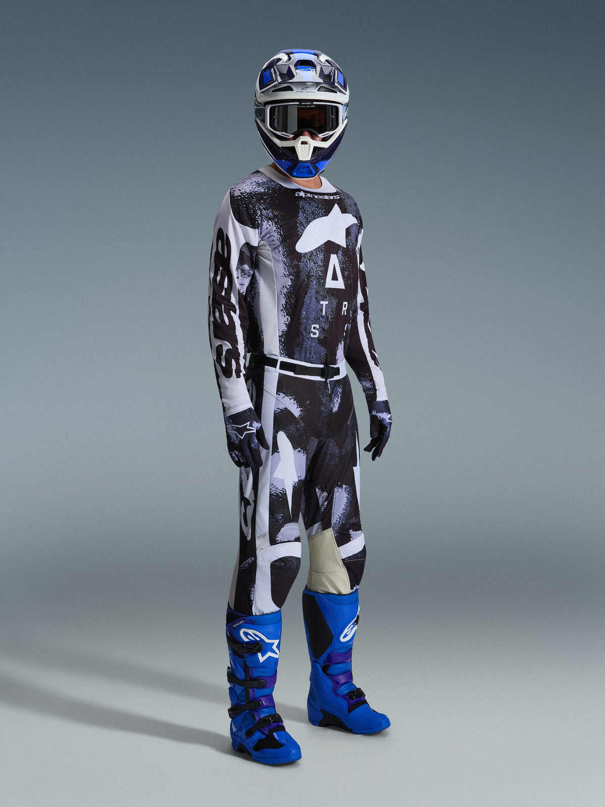 Alpinestars 2026 Racer Lahnd Gear Set, pilota di motocross con maglia e pantaloni a fantasia bianca e nera dal design astratto effetto invecchiato, grande logo Alpinestars bianco, stivali blu e viola vivaci e casco bianco e blu, outfit professionale completo per corse off-road.