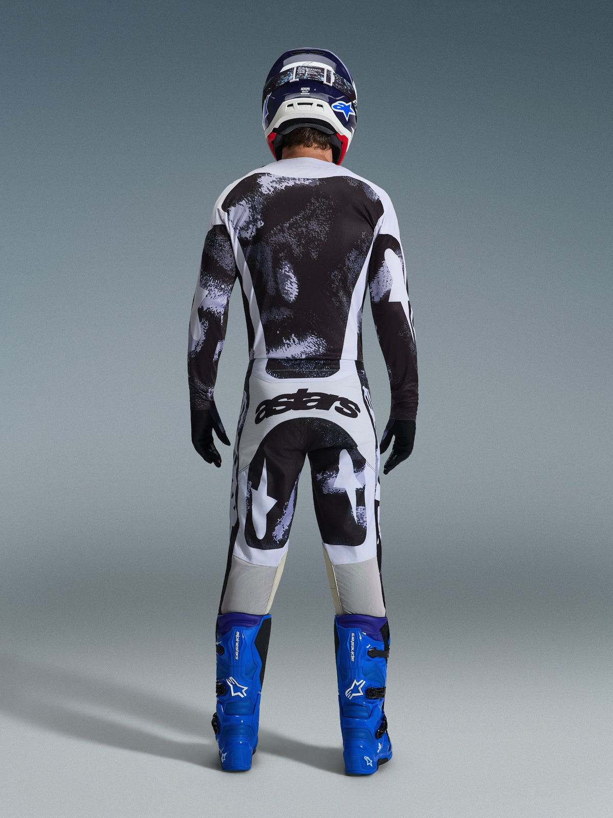 Pilota di motocross visto dal retro che indossa il completo Alpinestars 2026 Racer Lahnd, maglia e pantaloni con motivo astratto bianco e nero, logo 'astars' sulla seduta, abbinato a stivali blu e casco coordinato, abbigliamento professionale per corse fuoristrada.