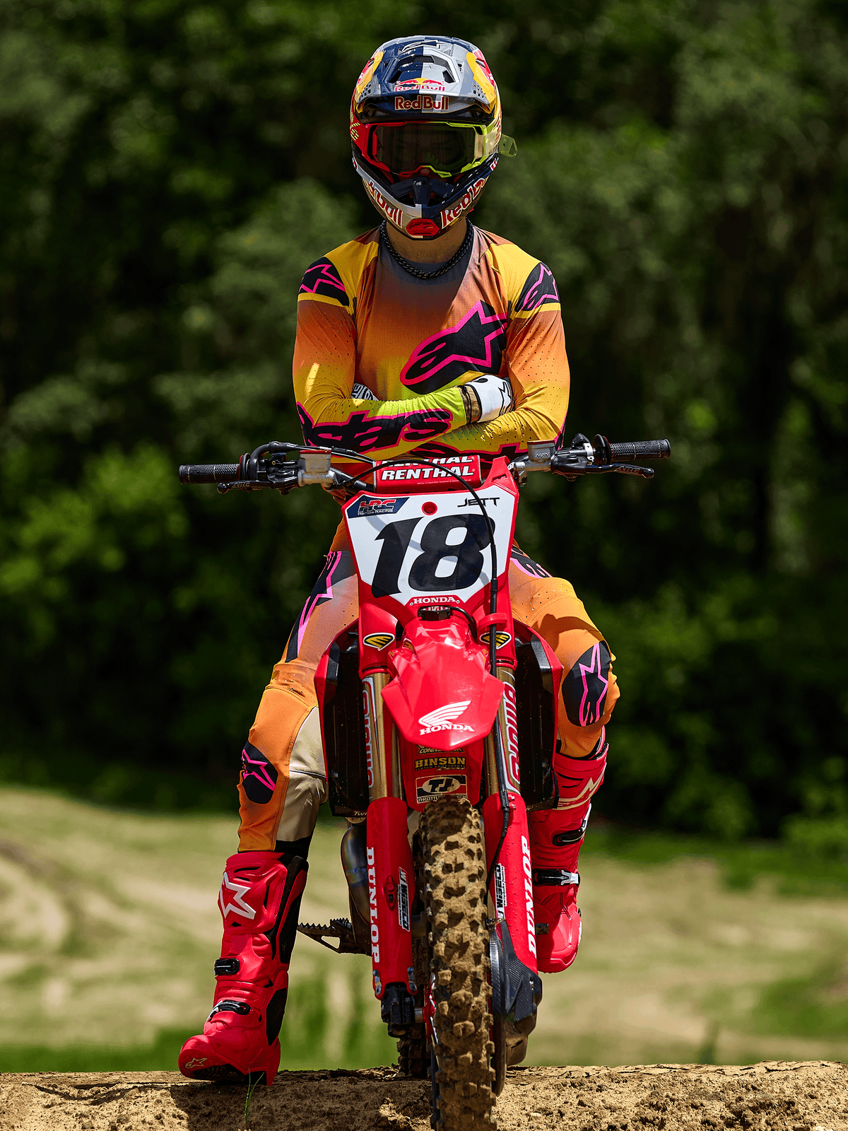 Pilota di motocross seduto su una moto da cross Honda rossa con il numero di gara 18, che indossa la maglia Alpinestars Supertech Lipan 2025 e i pantaloni coordinati in giallo e rosa, stivali Alpinestars rosa acceso, casco Red Bull, abbigliamento professionale da gara per prestazioni fuoristrada.