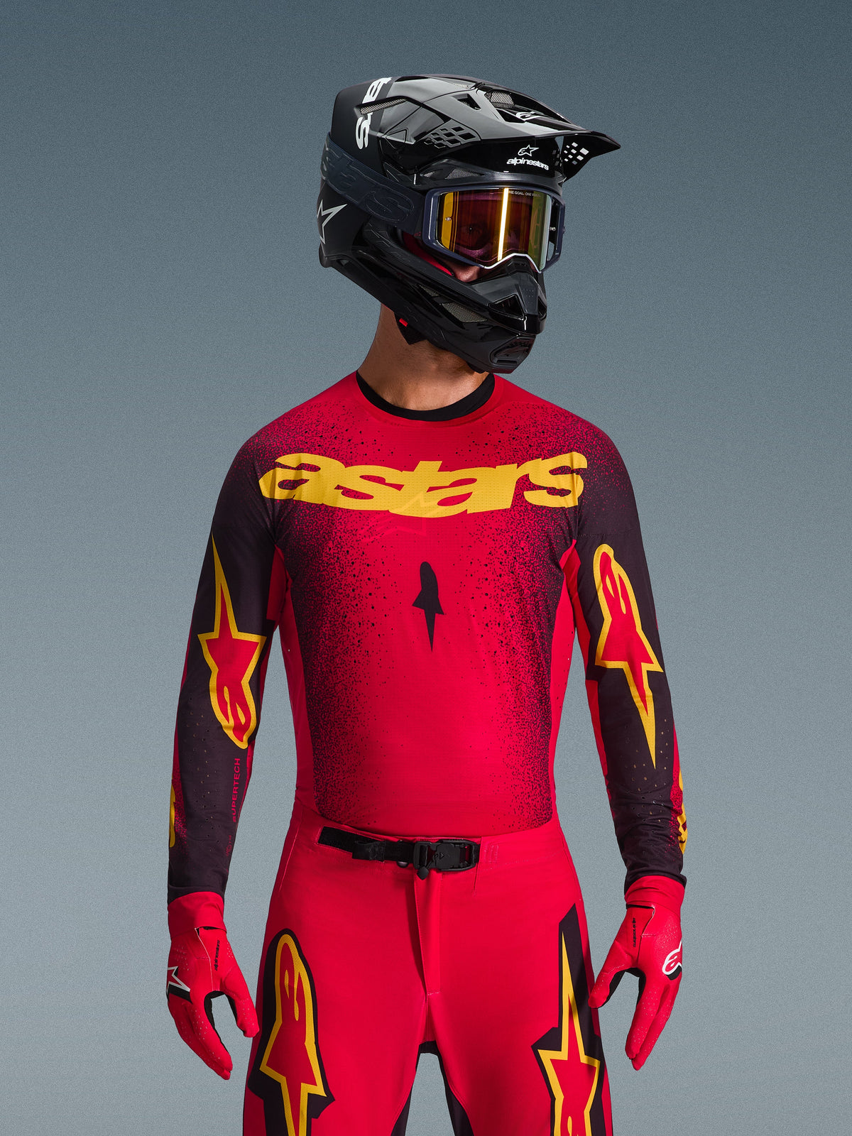 Alpinestars 2026 Supertech Scenz Gear Set, pilota di motocross con maglia rossa, marchio 'astars' giallo e dettagli neri picchiettati, pantaloni rossi con loghi a stella gialli sulle cosce, guanti da corsa rossi e casco nero con maschera gialla riflettente, completo da gara professionale per prestazioni fuoristrada