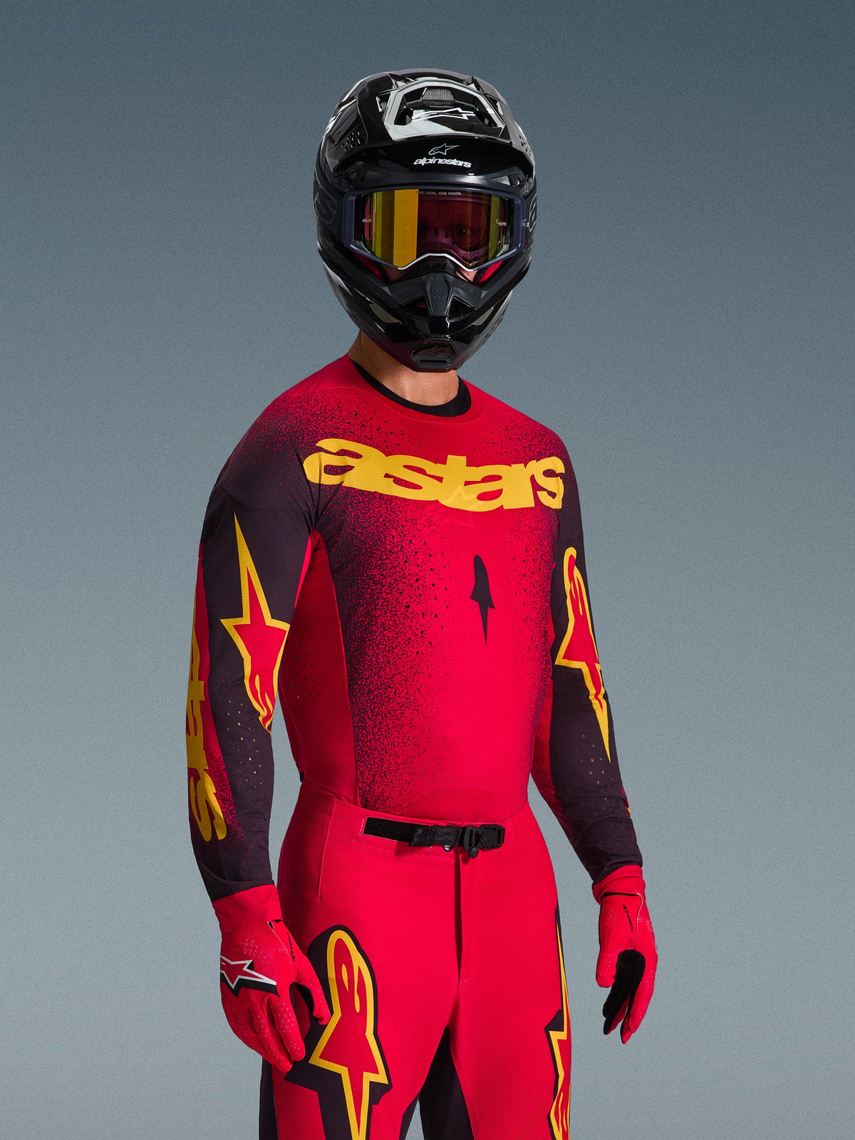 Pilota di motocross con completo Alpinestars Supertech Scenz 2026, maglia rossa e nera con logo 'astars' giallo e sfumatura punteggiata, pantaloni rossi coordinati, guanti rossi e casco nero con occhiali gialli, abbigliamento professionale da motocross
