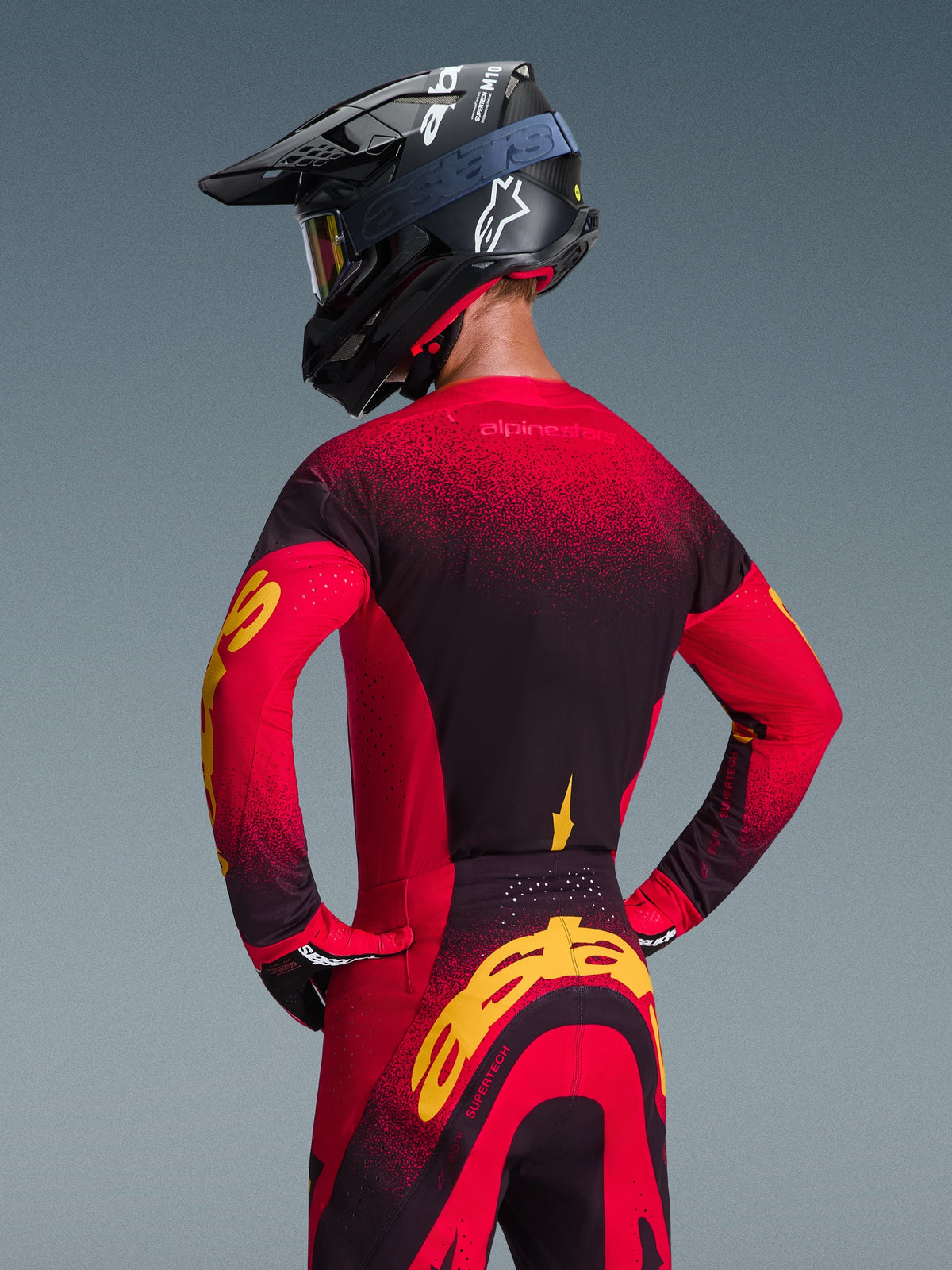 Pilota di motocross visto dal retro che indossa la maglia Alpinestars 2026 Supertech Scenz e i pantaloni coordinati, combinazione di colori rosso, nero e giallo, casco da gara nero con logo bianco, con branding giallo vistoso sulle maniche e sul retro, attrezzatura professionale da fuoristrada.