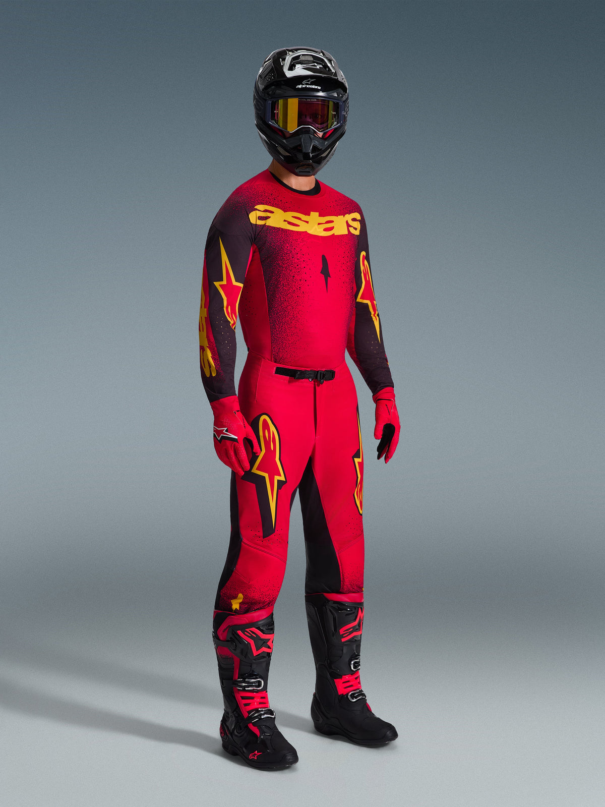 Pilota di motocross con abbigliamento completo da gara, maglia rossa con maniche nere e grafiche gialle, pantaloni rossi coordinati con pannelli neri e loghi gialli, casco nero con occhiali con lente gialla, stivali neri e rossi, guanti rossi e neri, completo Supertech Scenz 2026 coordinato per gare professionistiche di motocross