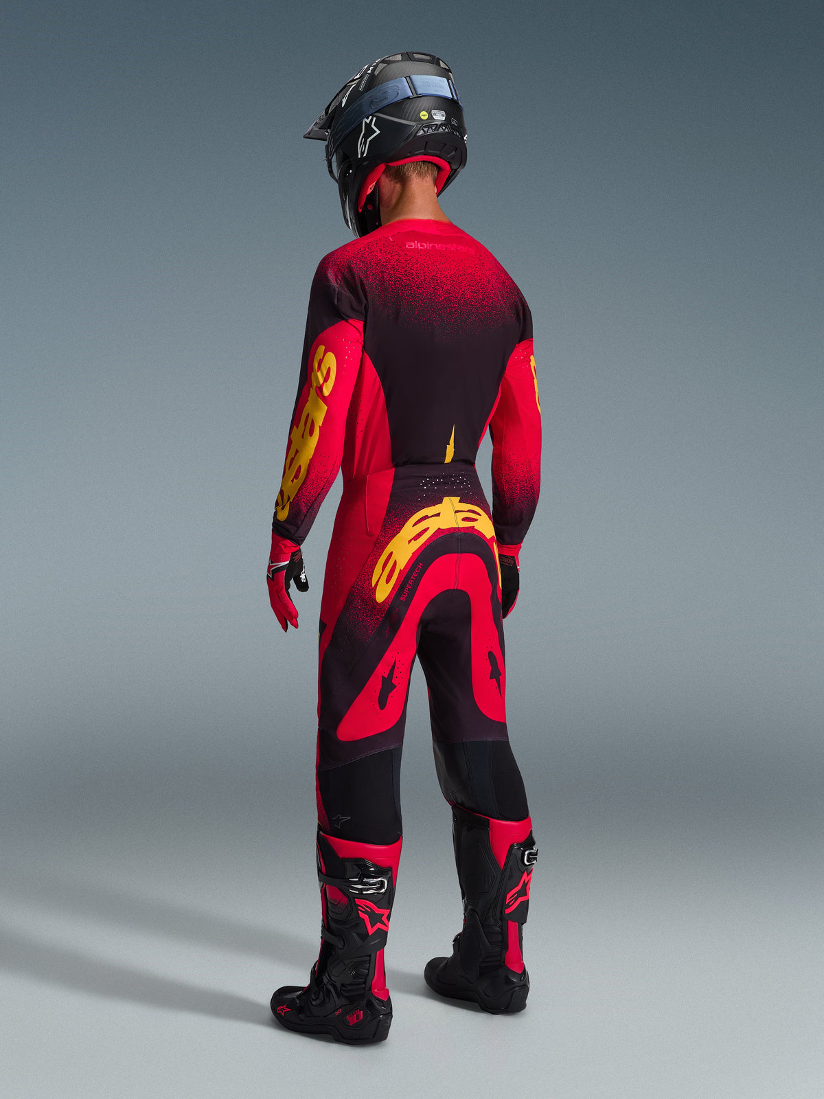 Set completo Alpinestars Supertech Scenz 2026, pilota di motocross visto da dietro, maglia rossa e nera con loghi gialli, pantaloni coordinati rossi e neri con logo giallo, guanti rossi, stivali da motocross neri e rossi e casco nero, abbigliamento da gara professionale coordinato.