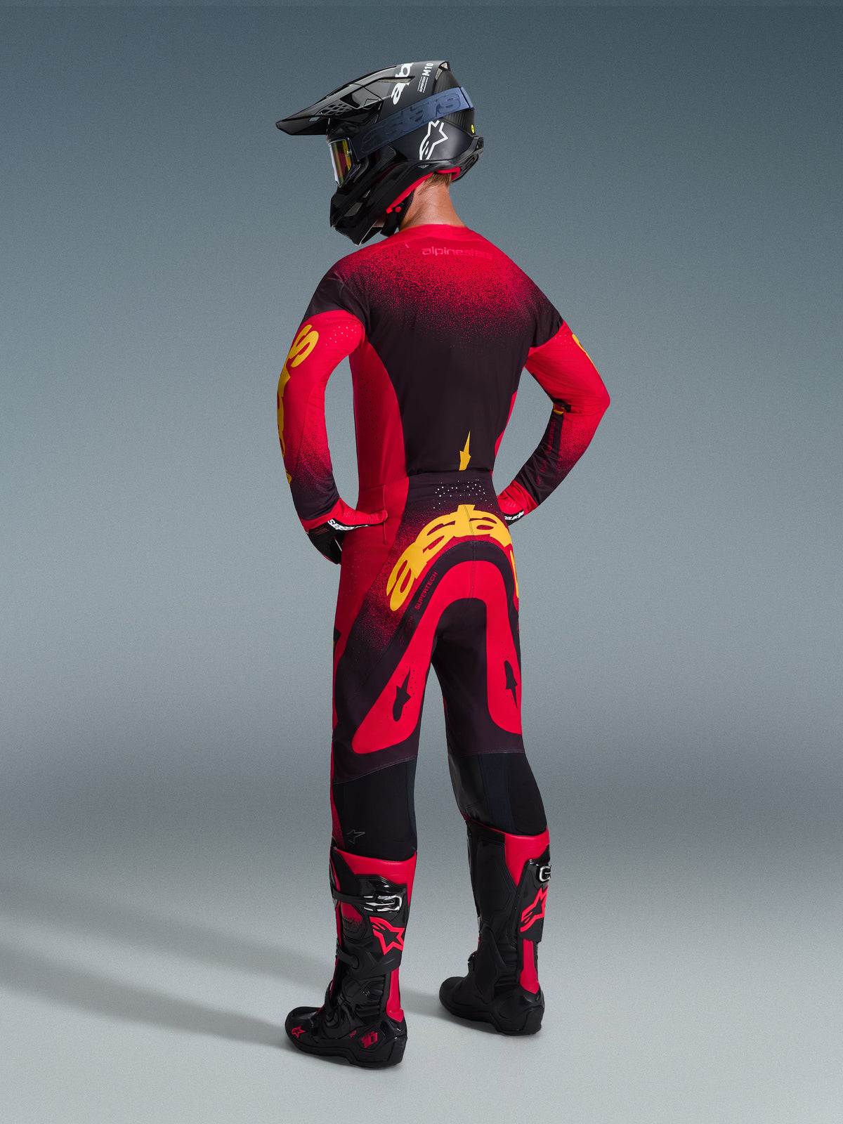Pilota di motocross con il completo Alpinestars Supertech Scenz 2026, maglia rossa e nera con accenti gialli, pantaloni coordinati con vistoso logo giallo sul retro, casco nero e stivali con dettagli rossi, outfit coordinato per gare professionistiche di motocross