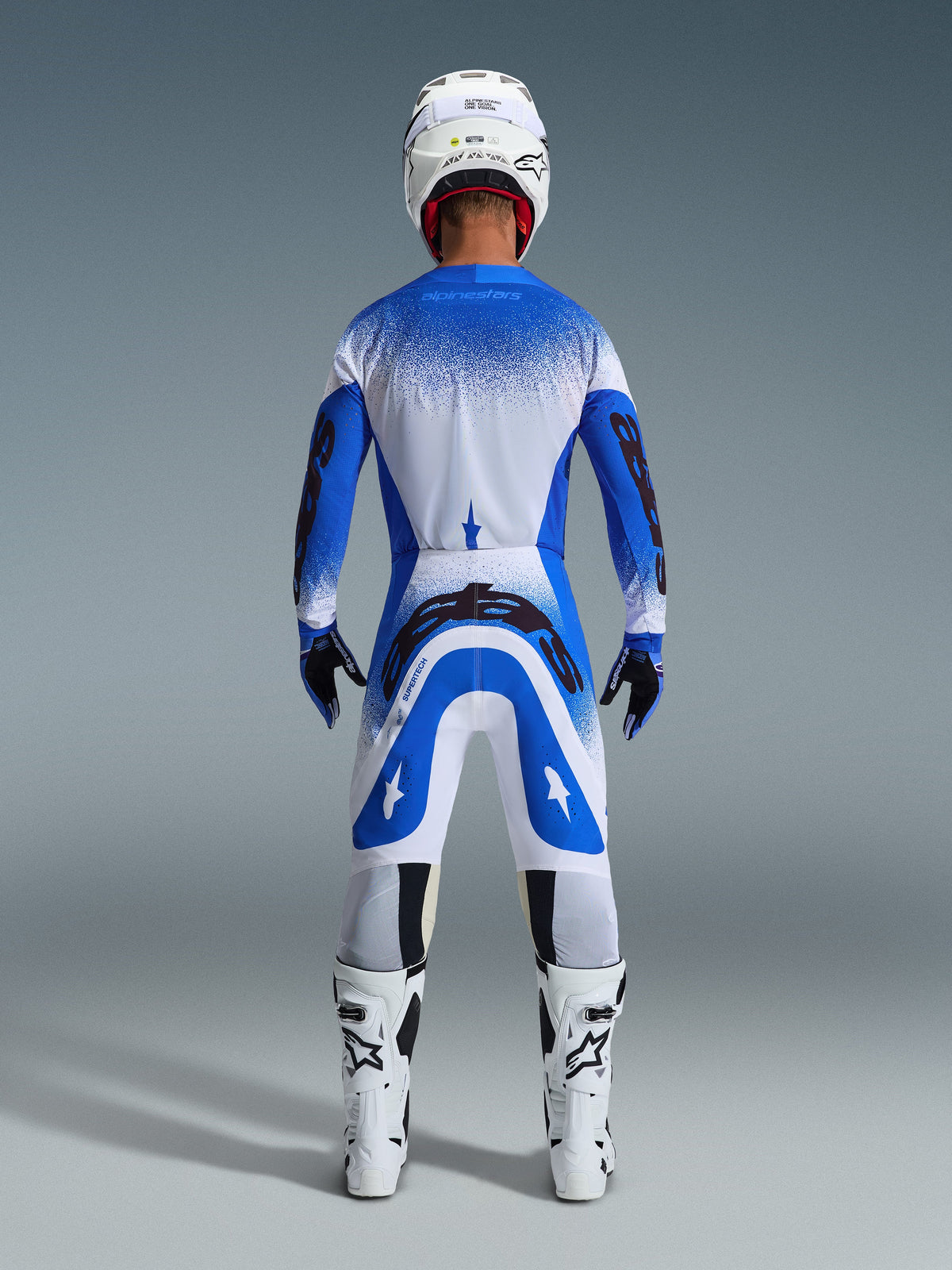 Pilota di motocross visto da dietro che indossa il completo Alpinestars Supertech Scenz 2026 in Ucla Blue White, maglia blu e bianca con marchio nero, pantaloni coordinati, casco bianco e stivali bianchi, equipaggiamento racing professionale per motocross fuoristrada