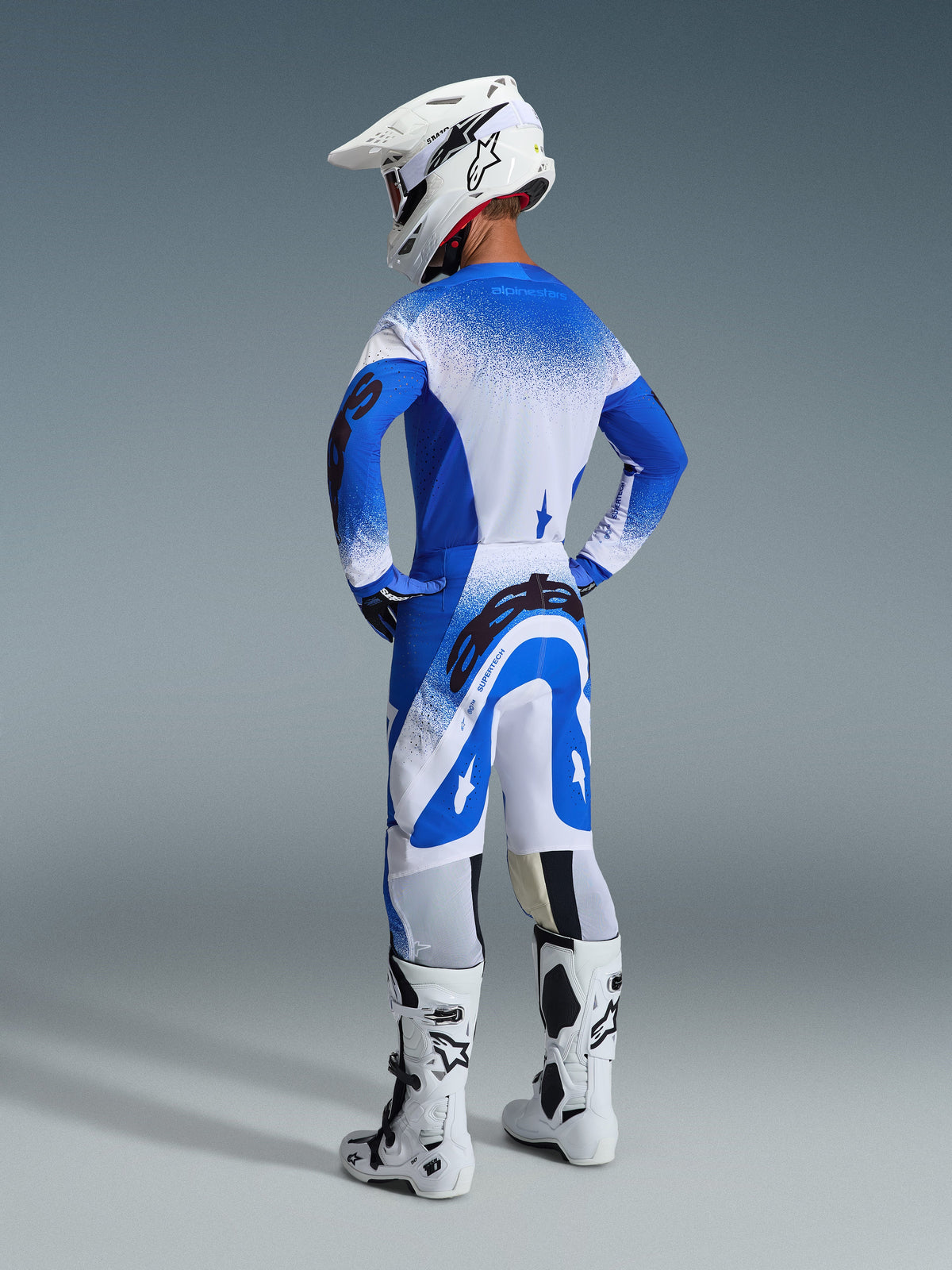 Alpinestars 2026 Supertech Scenz Gear Set, vista posteriore di un pilota che indossa maglia e pantaloni Ucla Blue White, casco da motocross bianco e stivali da gara bianchi, completo da gara di livello professionale