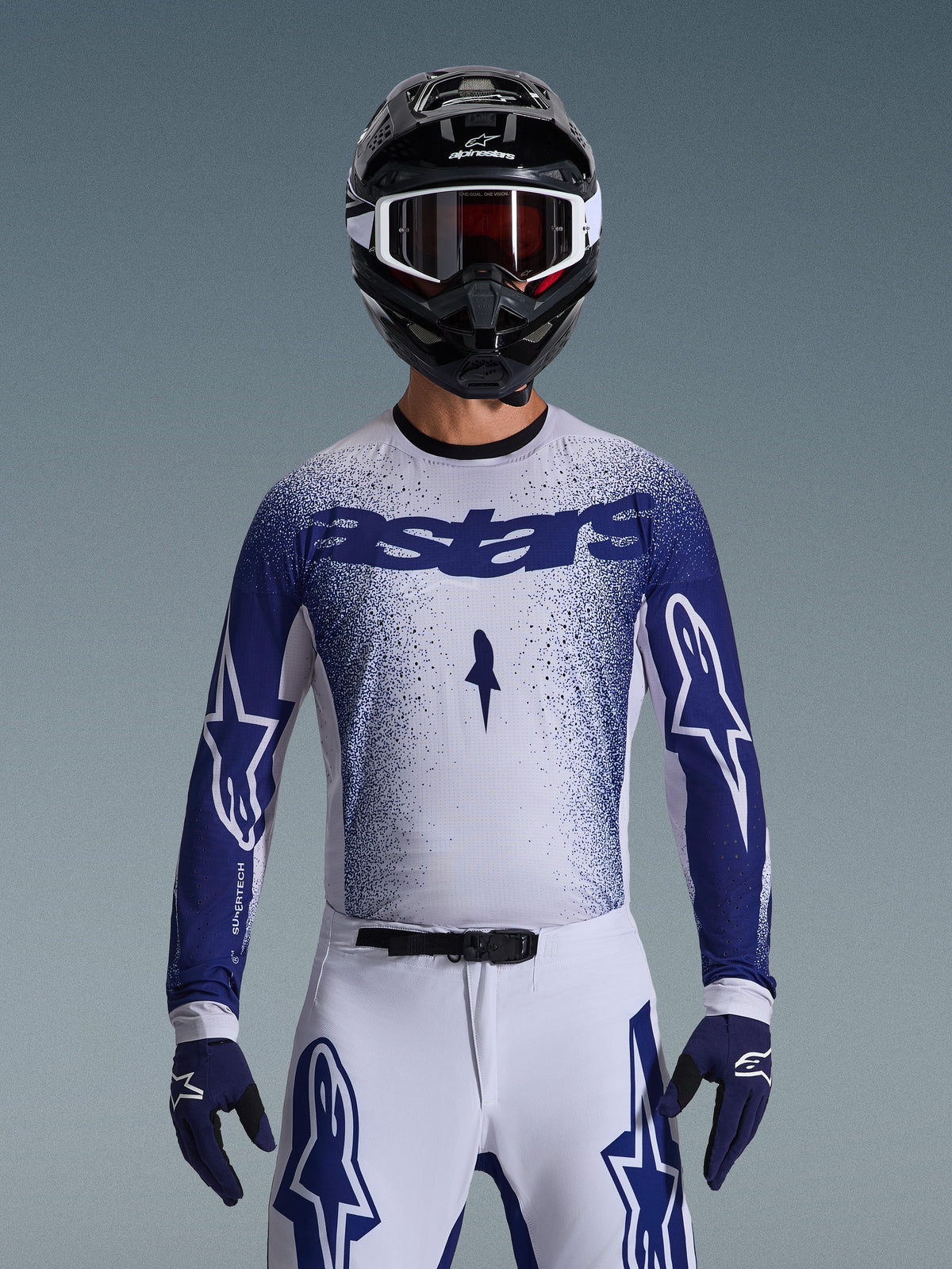 Pilota di motocross con maglia Alpinestars Supertech Scenz 2026, grigio chiaro e blu scuro con design a sfumatura puntinata e logo 'astars' in evidenza. Indossata con pantaloni da motocross bianchi coordinati, guanti blu scuro e casco nero con maschera, completo per gare professionistiche di fuoristrada.