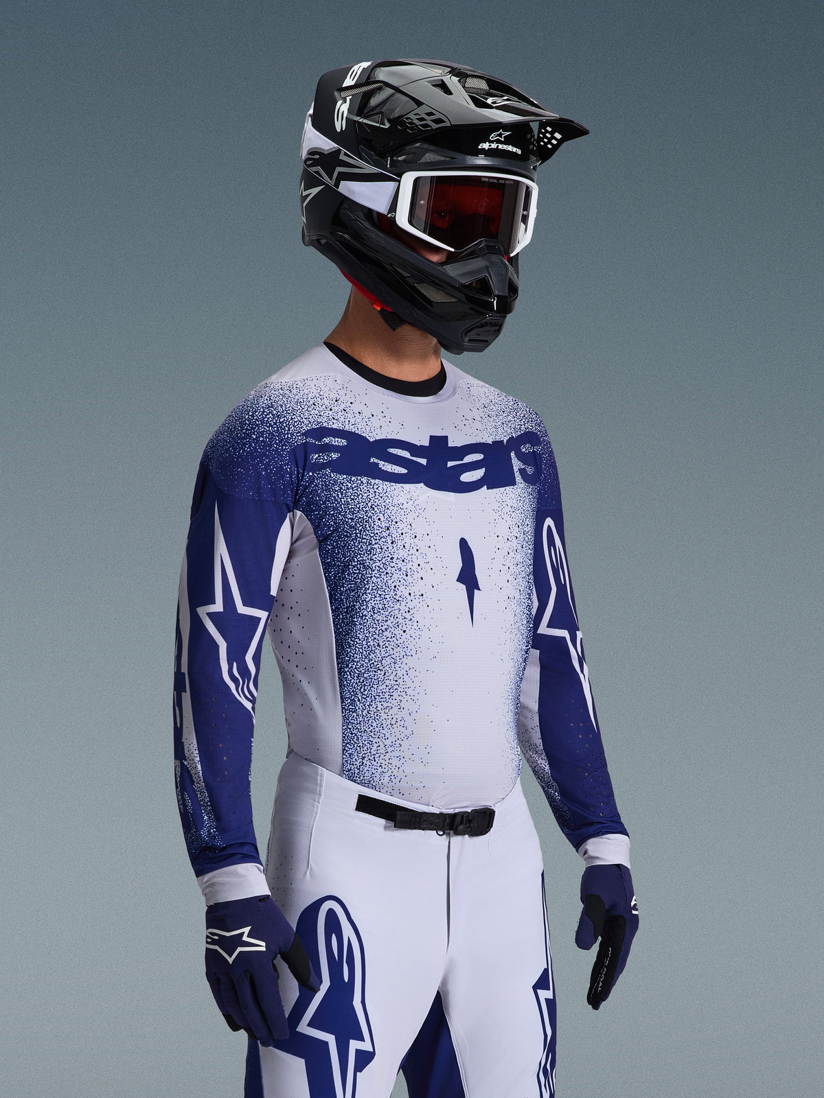 Pilota di motocross che indossa la maglia Supertech Scenz 2026 in grigio chiaro e blu scuro, abbinata a pantaloni da gara coordinati bianchi e blu, guanti blu e un casco nero con maschera bianca. La maglia presenta un effetto sfumato blu puntinato su petto e maniche, vestibilità atletica professionale e il marchio 'astars' in evidenza.