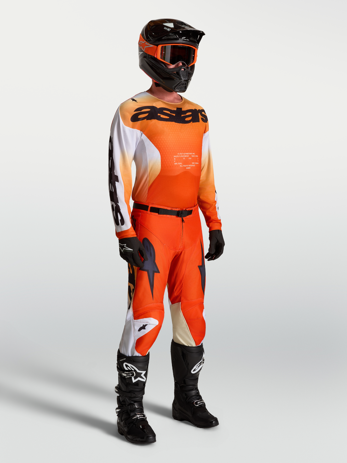 Alpinestars Maglia Racer Amber, maglia da motocross da uomo, arancione e nero, in tessuto tecnico leggero, indossata da un modello con equipaggiamento completo da gara e casco.