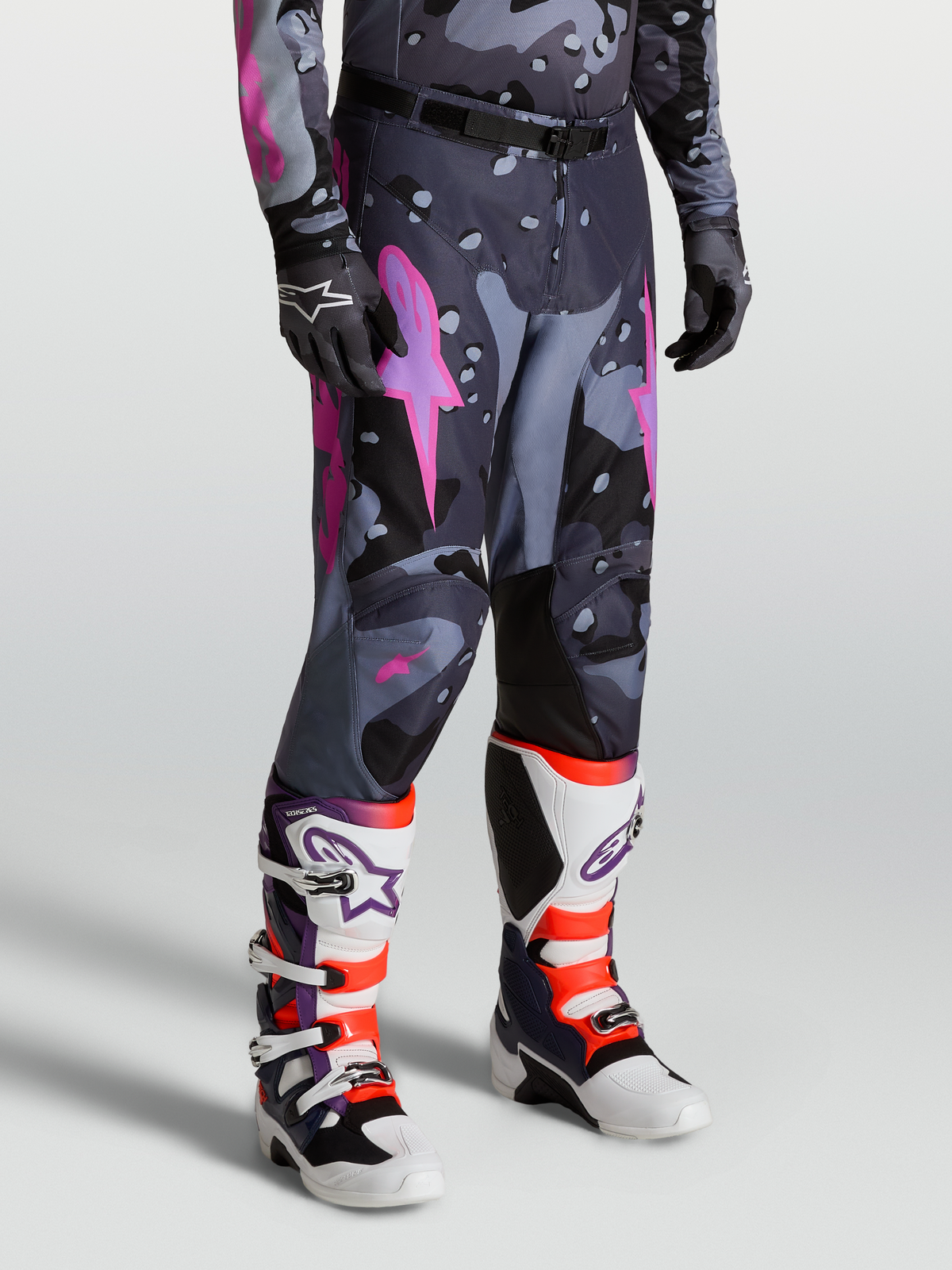 Alpinestars Pantaloni Racer Lone, pantaloni motocross da uomo, grigio camo e viola, design ergonomico precurvato e rinforzi in poliestere 600D, indossati con stivali Tech 10 bianchi e viola.