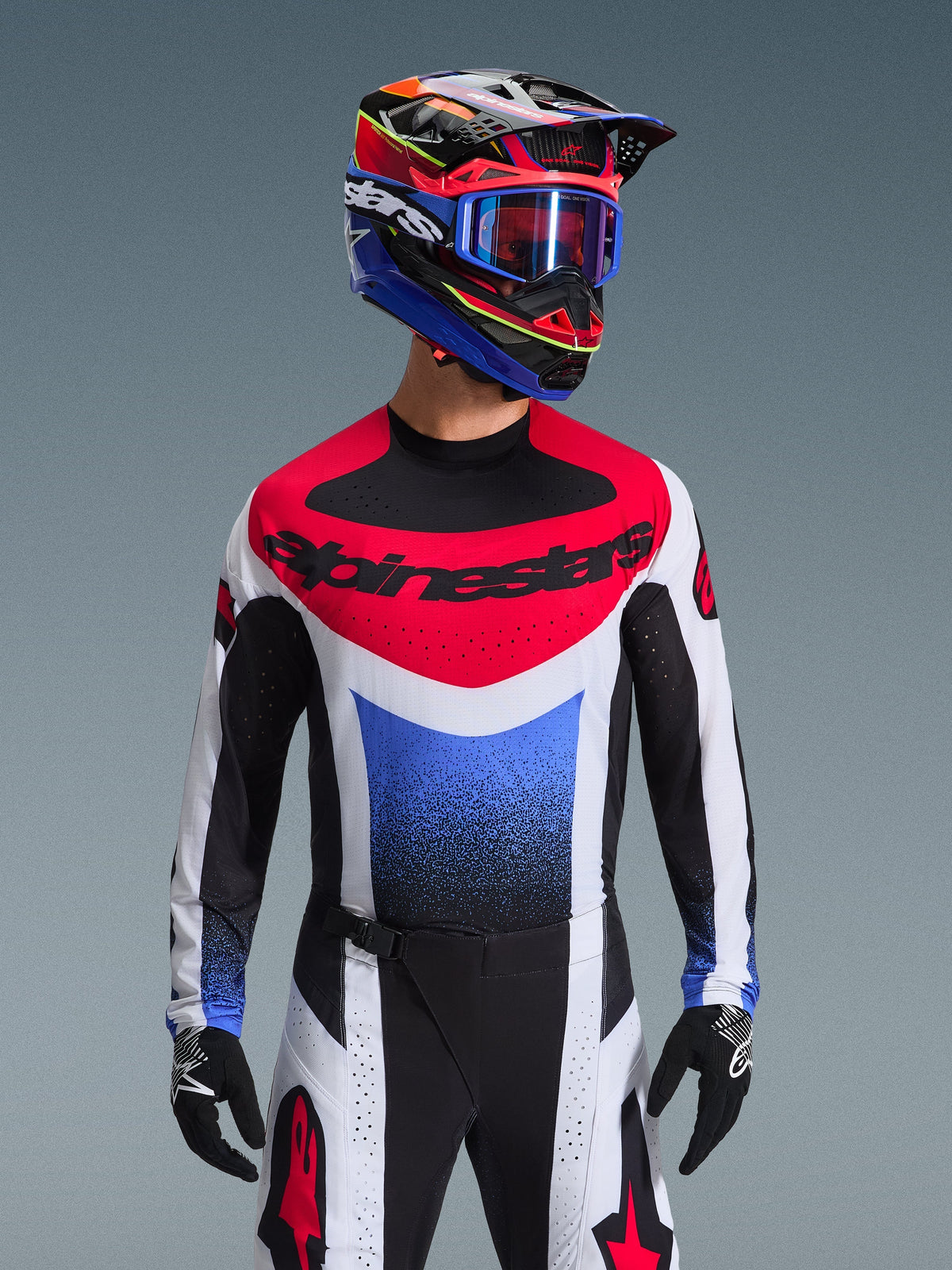 Alpinestars 2026 Techstar Knif Jersey, maglia da motocross, rosso, bianco e blu, con un vistoso logo sul petto e busto sfumato, indossata da un pilota con casco multicolore coordinato, occhiali blu e guanti neri, abbigliamento professionale da fuoristrada.