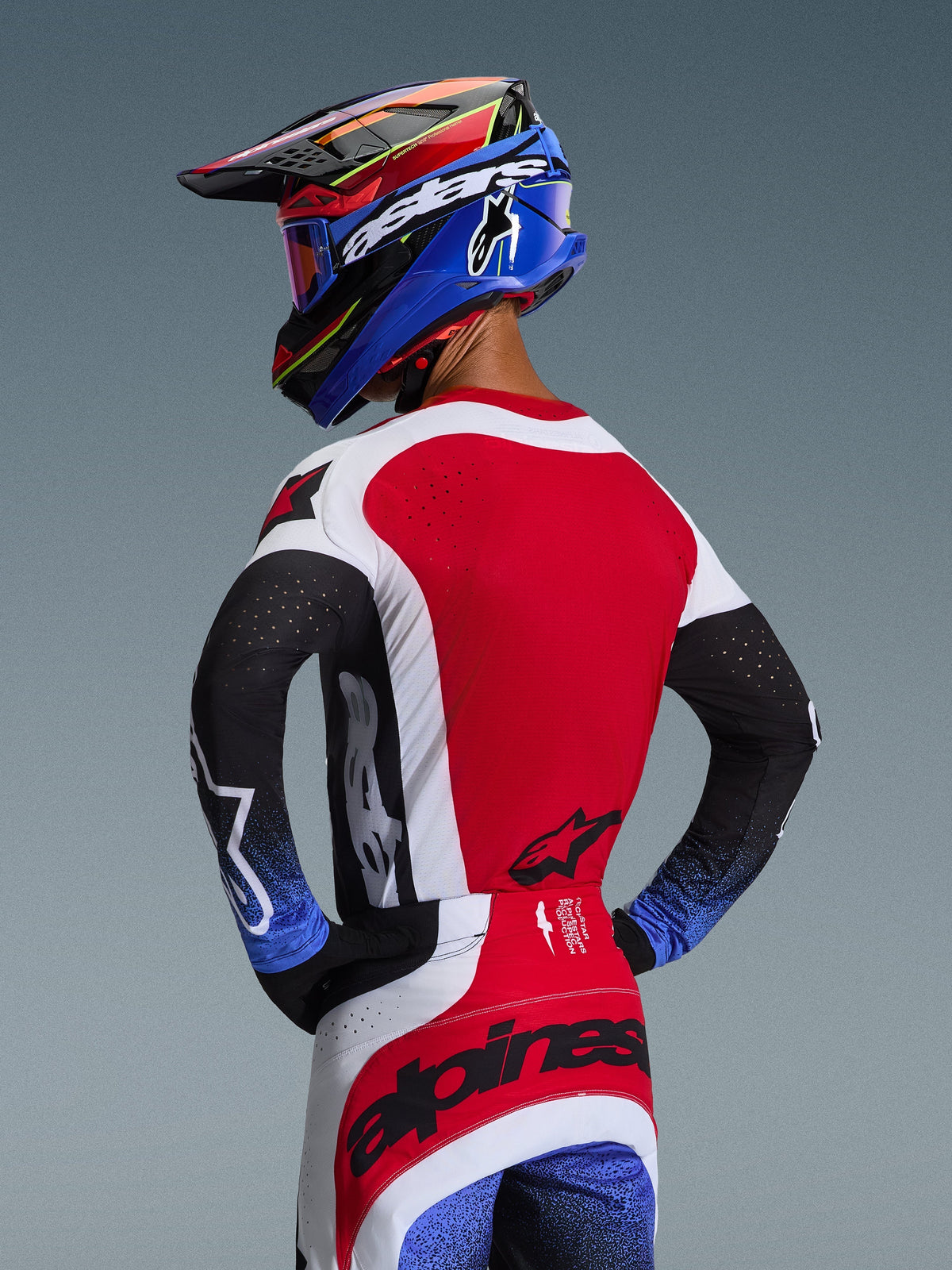 Pilota di motocross visto da dietro che indossa la maglia Techstar Knif 2026 in rosso, bianco e nero, con perforazioni tagliate al laser sulla schiena e branding evidente, abbinata a pantaloni coordinati con accenti blu e un casco Alpinestars blu e nero, estetica racing professionale fuoristrada