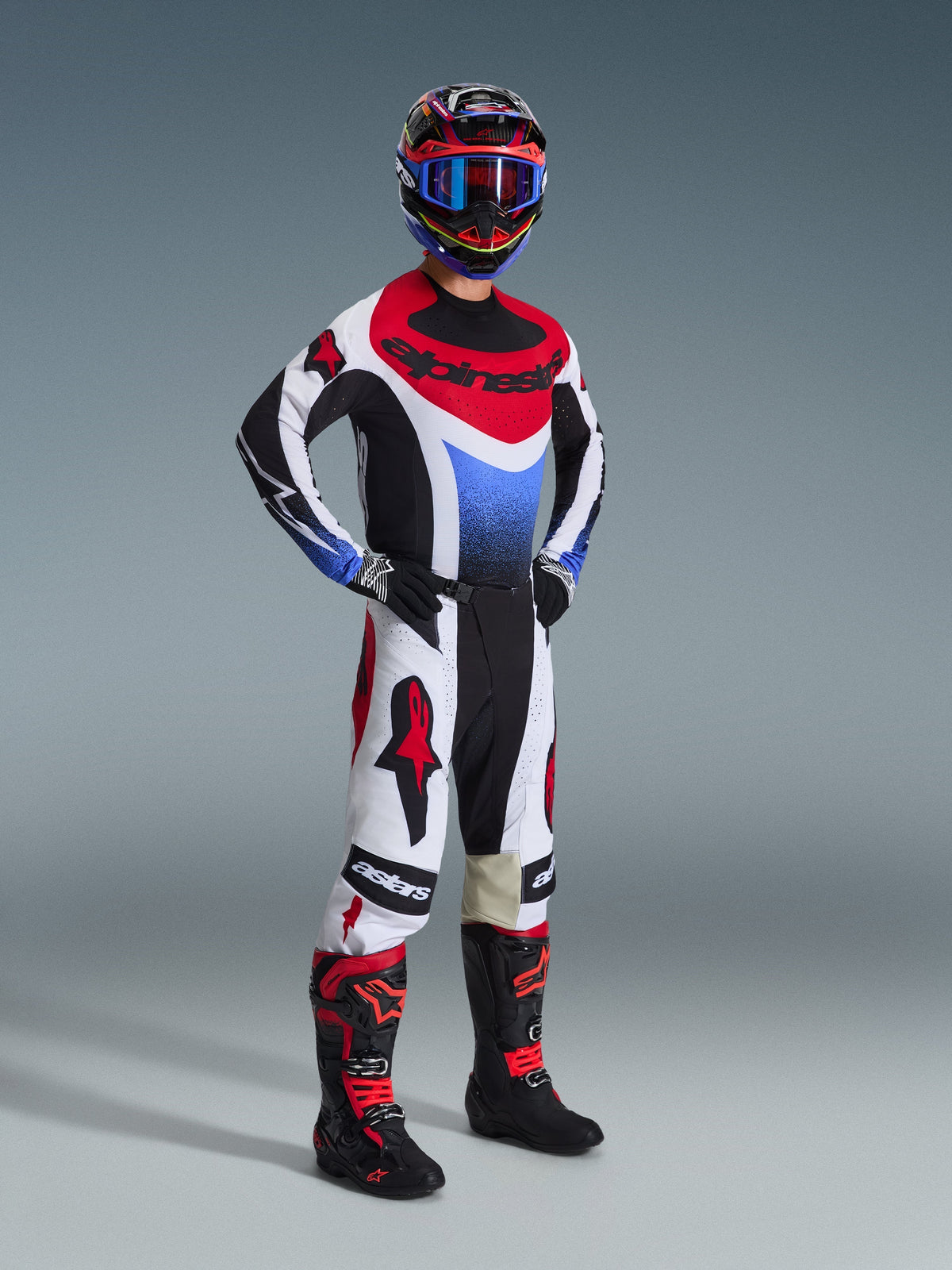 Pilota di motocross che indossa la maglia Alpinestars Techstar Knif 2026 in nero, bianco e rosso con accenti blu, abbinata a pantaloni bianchi e rossi coordinati, stivali neri e rossi e casco con occhiali blu, kit completo da gara off-road