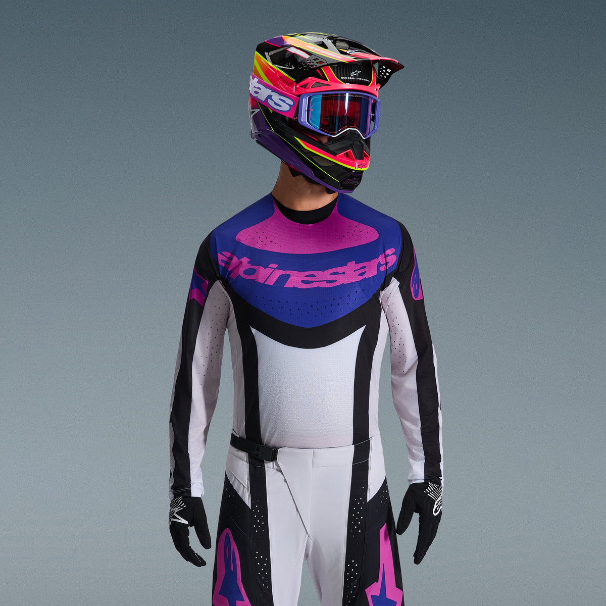 Pilota di motocross con completo Alpinestars Techstar Knif 2026, maglia con grafica blu e rosa sul petto e busto bianco, pantaloni bianchi con pannelli neri e loghi rosa, casco multicolore viola e rosa con occhiali coordinati, guanti neri, abbigliamento professionale per gare fuoristrada